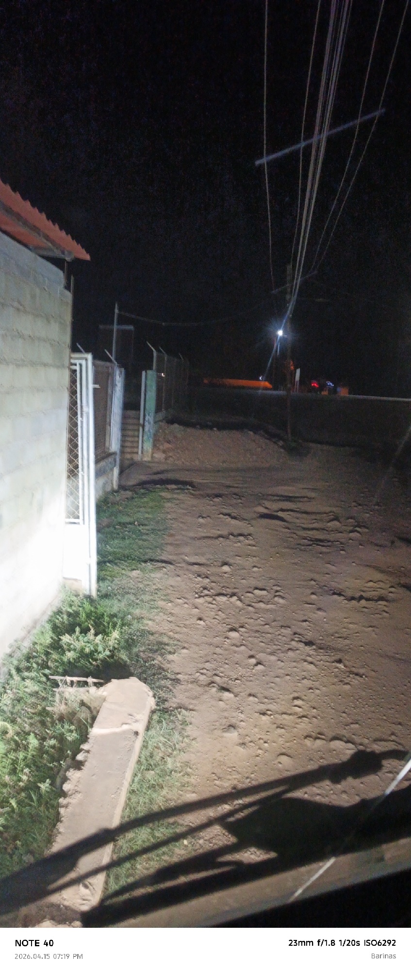 Captured at night in Barinas, Venezuela, the image shows a dirt road illuminated by an unseen light source, possibly a vehicle. To the left is a concrete building with a red roof and a fence, alongside vegetation. Across the road is another fence, beyond which are distant lights, possibly from a vehicle. Overhead, power lines stretch across the frame. The photo was taken on April 15, 2026, at 7:19 PM. The camera settings were 23mm, f/1.8, 1/20s, ISO 6292.
