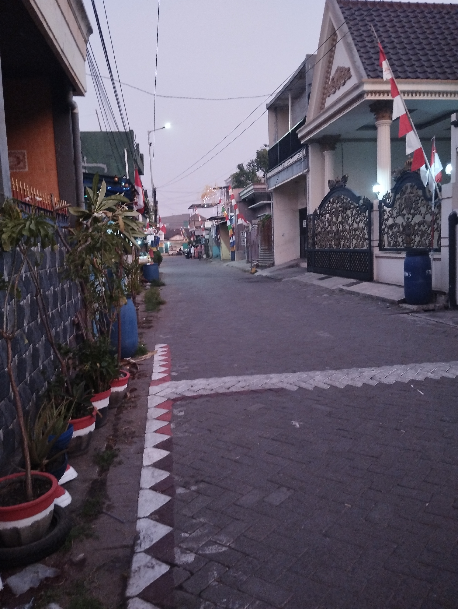 The image captures a residential street in Kepuh Kiriman, Indonesia, during what appears to be twilight. The scene is characterized by a paved road with a decorative border of red and white triangular patterns on the left side. Along the left edge of the road, a wall is visible, covered in a textured, dark gray material. Several potted plants are arranged along this wall, some with red and white painted rims.
On the right side of the street, a house with a prominent gabled roof stands, decorated with ornate carvings under the eaves. A large, decorative black gate with intricate metalwork leads to the property. Flags, red and white, are displayed prominently on the house and along the street, suggesting a national celebration or event.
Further down the street, more houses and flags are visible, indicating a densely populated residential area. A few people are present in the distance, walking along the street. Large blue barrels, likely for water storage, are placed near the gate and further down the road. The sky is a pale, overcast hue, common during dusk or dawn. Overhead, a network of electrical wires crisscrosses the sky, a common sight in urban and suburban Indonesian settings. The overall impression is of a quiet, community-oriented street decorated for a special occasion.