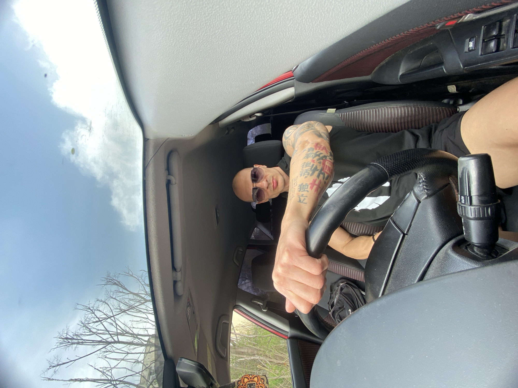 A man with a shaved head and sunglasses is driving a car. His left arm is tattooed with Asian characters. The view through the windshield shows bare trees and a cloudy sky. The car's interior is visible, including the steering wheel, dashboard, and part of the passenger seat. The location is not explicitly stated as Pleven, Bulgaria, or any specific city.