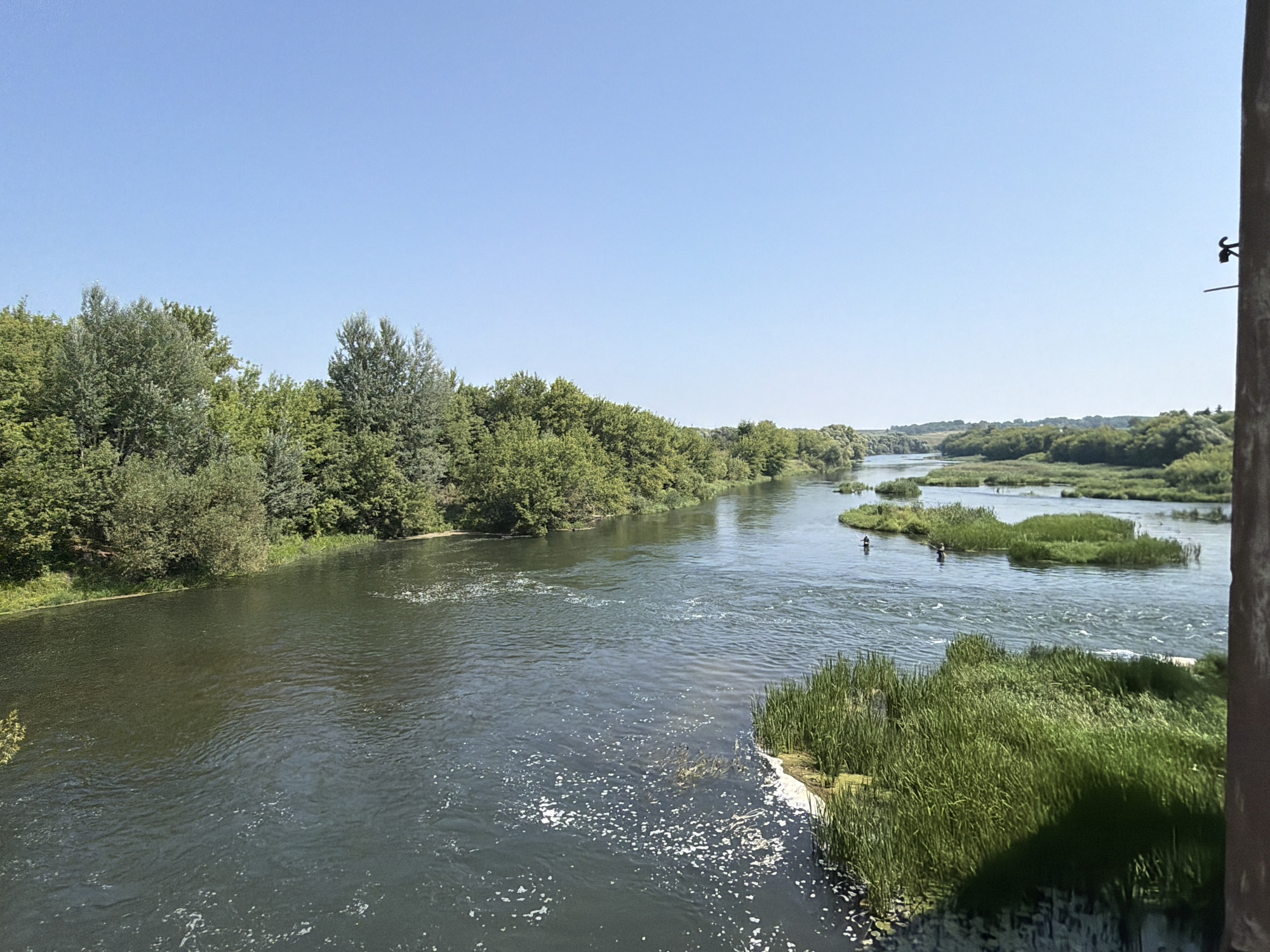 The image depicts a wide, tranquil river with lush green trees lining its banks. The sky is a clear, bright blue. There are small islands of reeds and vegetation in the river, and a few people are visible fishing or boating. The scene is set in a natural, rural environment, possibly a park or natural reserve within the vicinity of Троекурово, Russia.