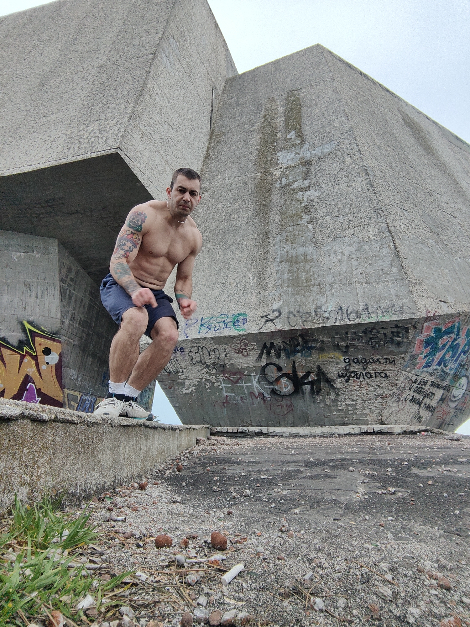 A man is standing on a concrete ledge in front of a large concrete structure with graffiti. The man is shirtless and wearing shorts, socks and sneakers. He has tattoos on his left arm and is clenching his fists, looking directly at the camera. The structure behind him is large and angular, seemingly a monument or building of Brutalist architecture. The surrounding area is rough, with broken bricks and weeds visible. The photo was taken at Buzludzha Monument in Varna, Bulgaria.