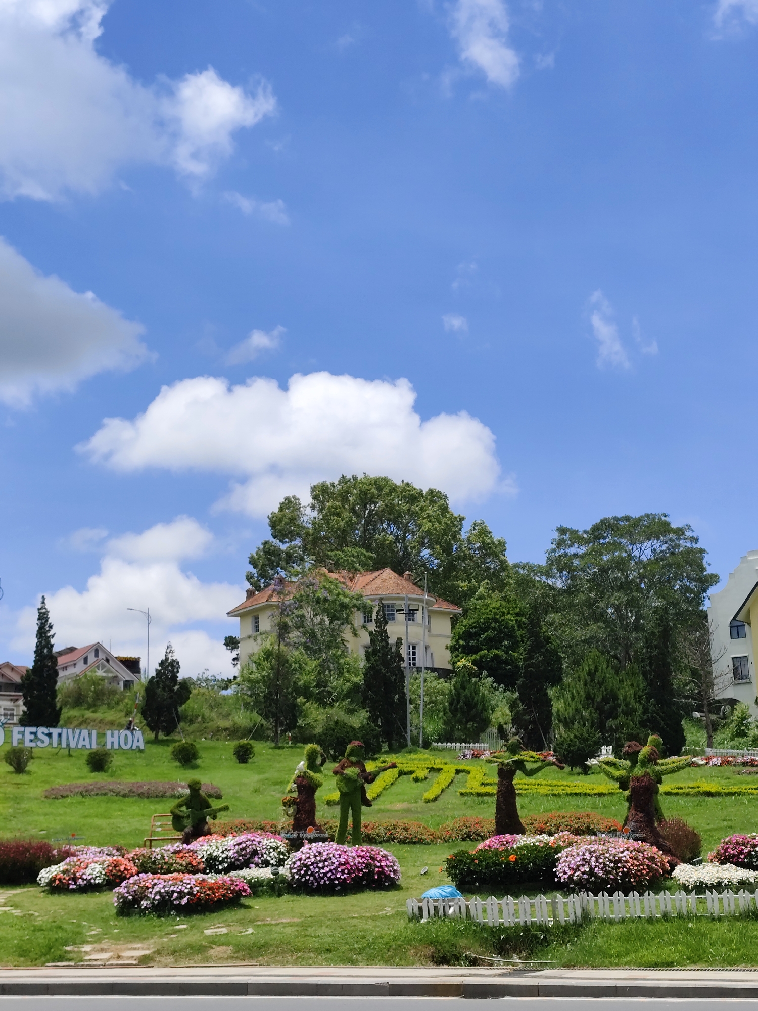 A wide shot shows a landscaped garden in the city of Da Lat, Vietnam, under a partly cloudy sky. A large flower bed in the foreground is filled with multicolored flowers, bordered by a low white picket fence. Behind the flower bed, green sculptures of musicians play on a grassy hill. To the left of the musicians, a large sign says "D FESTIVAL HOA." In the background, several buildings and trees are visible. The sky is a bright blue with scattered white clouds. The scene is shot from a slightly elevated position, with a road or sidewalk visible at the bottom of the frame.