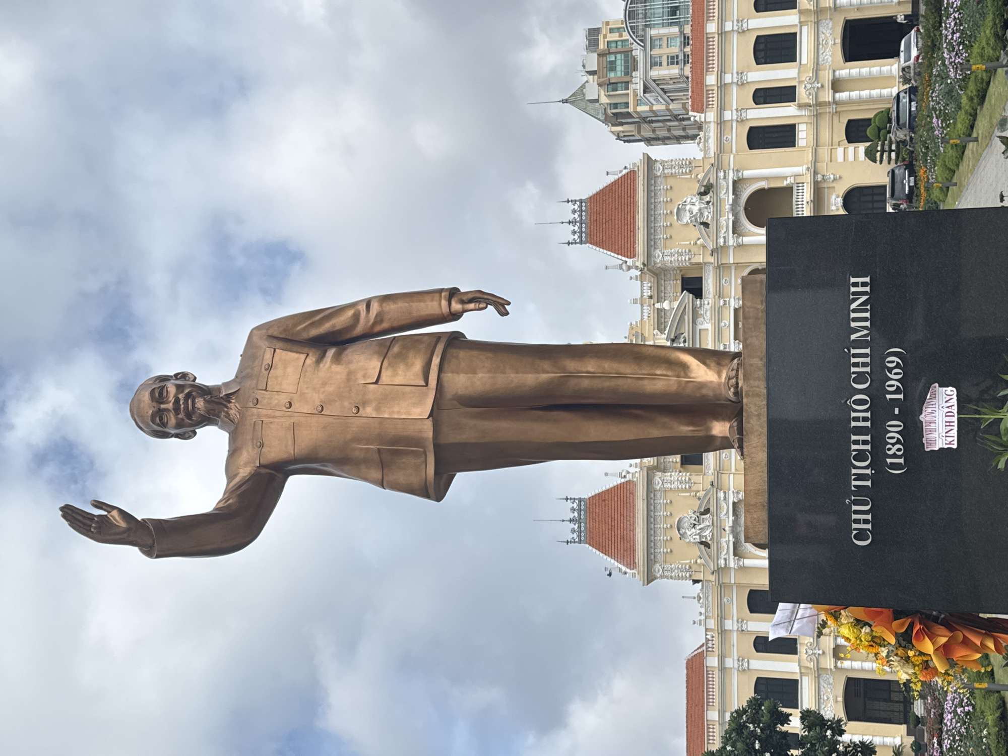 A full-body bronze statue of Ho Chi Minh, wearing a suit and with his right arm raised in a wave, stands on a black pedestal in front of a large, ornate yellow building. The building has a red-tiled roof, arched windows, and classical architectural details. In the foreground, to the left of the pedestal, is a bouquet of yellow and orange flowers. The sky is cloudy. The text on the pedestal reads "CHỦ TỊCH HỒ CHÍ MINH (1890 - 1969)". The scene is located in Ho Chi Minh City, Vietnam.