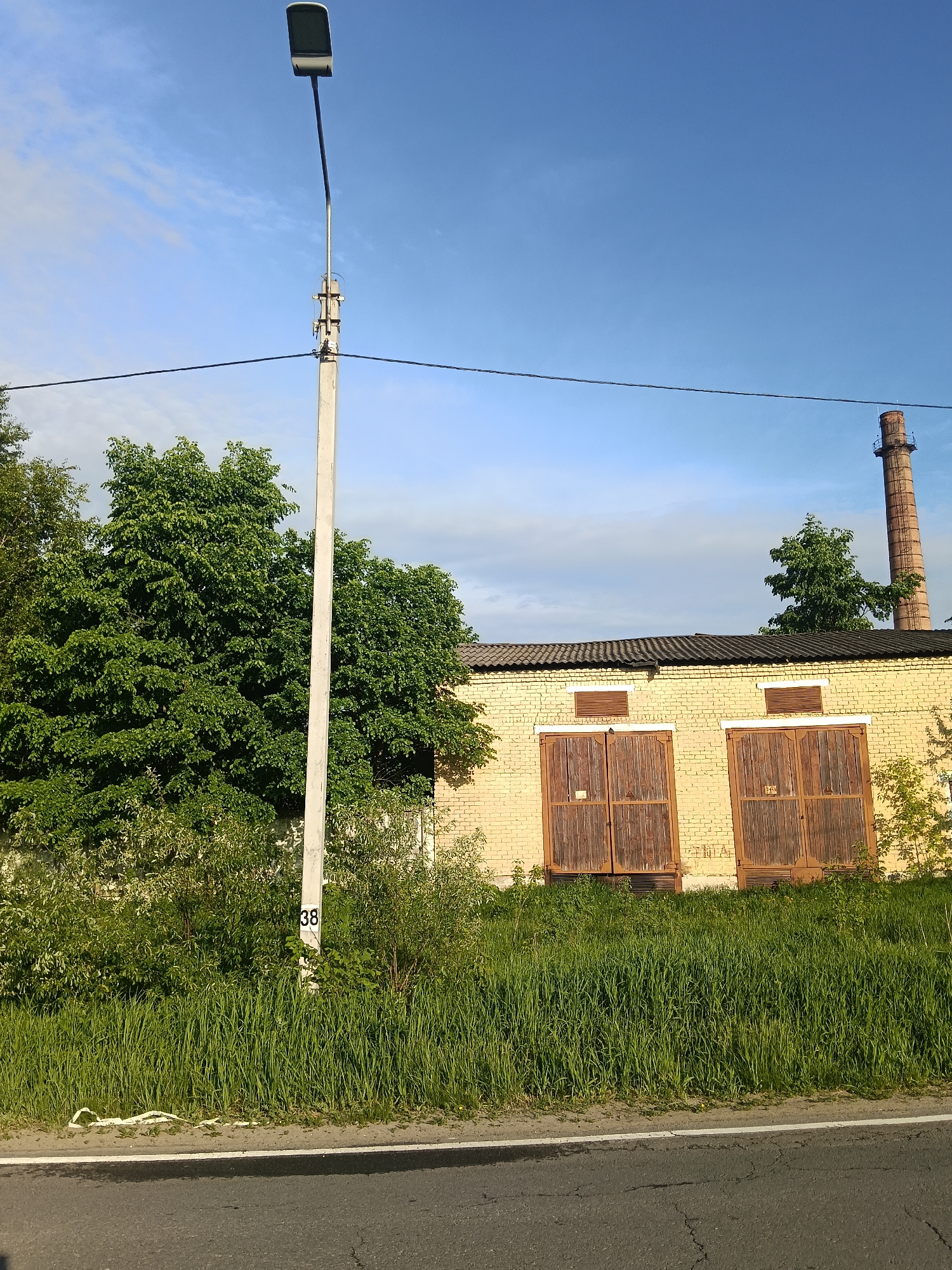 The photograph depicts an outdoor scene in Красный Маяк, Russia, featuring a light yellow brick building with two large, weathered, brown wooden doors. Above the building is a tall, slender, brick smokestack. A light pole stands in front of the building. The environment is overgrown with grass and foliage. A road is in the foreground. The sky is blue with scattered clouds. A power line runs horizontally across the top of the image. A sign with the number "38" is visible near the base of the light pole.