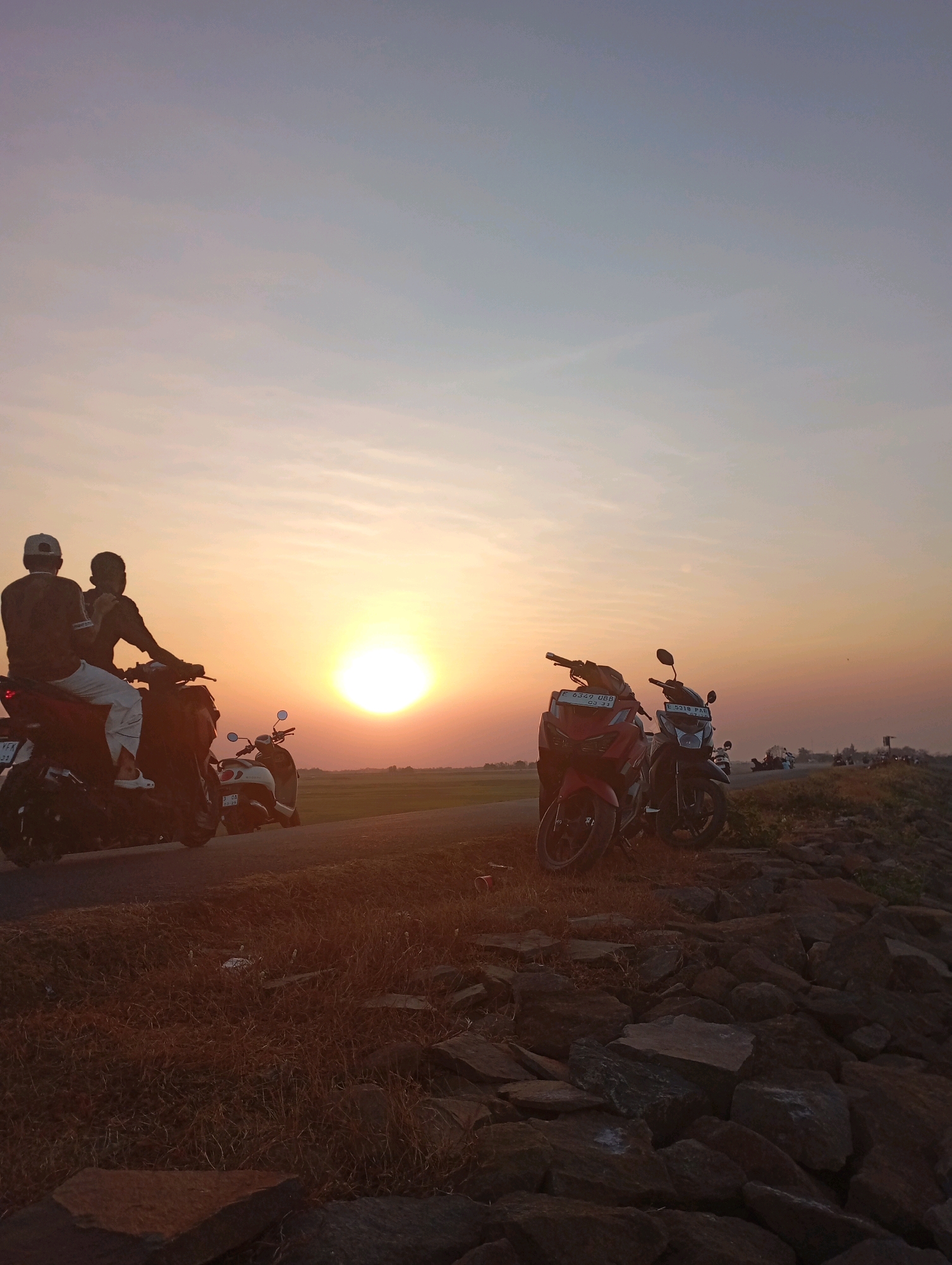 Two individuals are seated on a scooter, viewed from behind, facing a sunset. The sky exhibits a gradient of colors, from pale blue at the top to hues of orange and yellow near the horizon. A bright, circular sun is positioned low in the sky, casting a warm glow. In the foreground, several other motorcycles are parked. The ground is covered with dry grass and scattered rocks, creating a textured surface. The scene appears to be an open area, possibly a roadside or a field, with hints of a rural landscape in the background. The location is identified as Haurkolot, Indonesia.