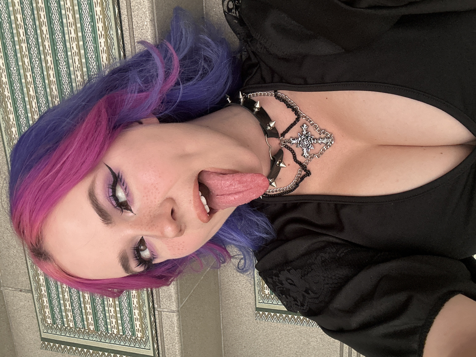 The image is a close-up, angled selfie of a young woman with blue and pink dyed hair. She is wearing a black top and a spiked collar with a chain necklace featuring a floral cross pendant. She is sticking her tongue out. Her makeup includes purple eyeshadow and winged eyeliner. She appears to be lying on a patterned surface, possibly a tiled floor or rug. Although the prompt mentions City of Zagreb, Croatia, there is no visible evidence to confirm this location.