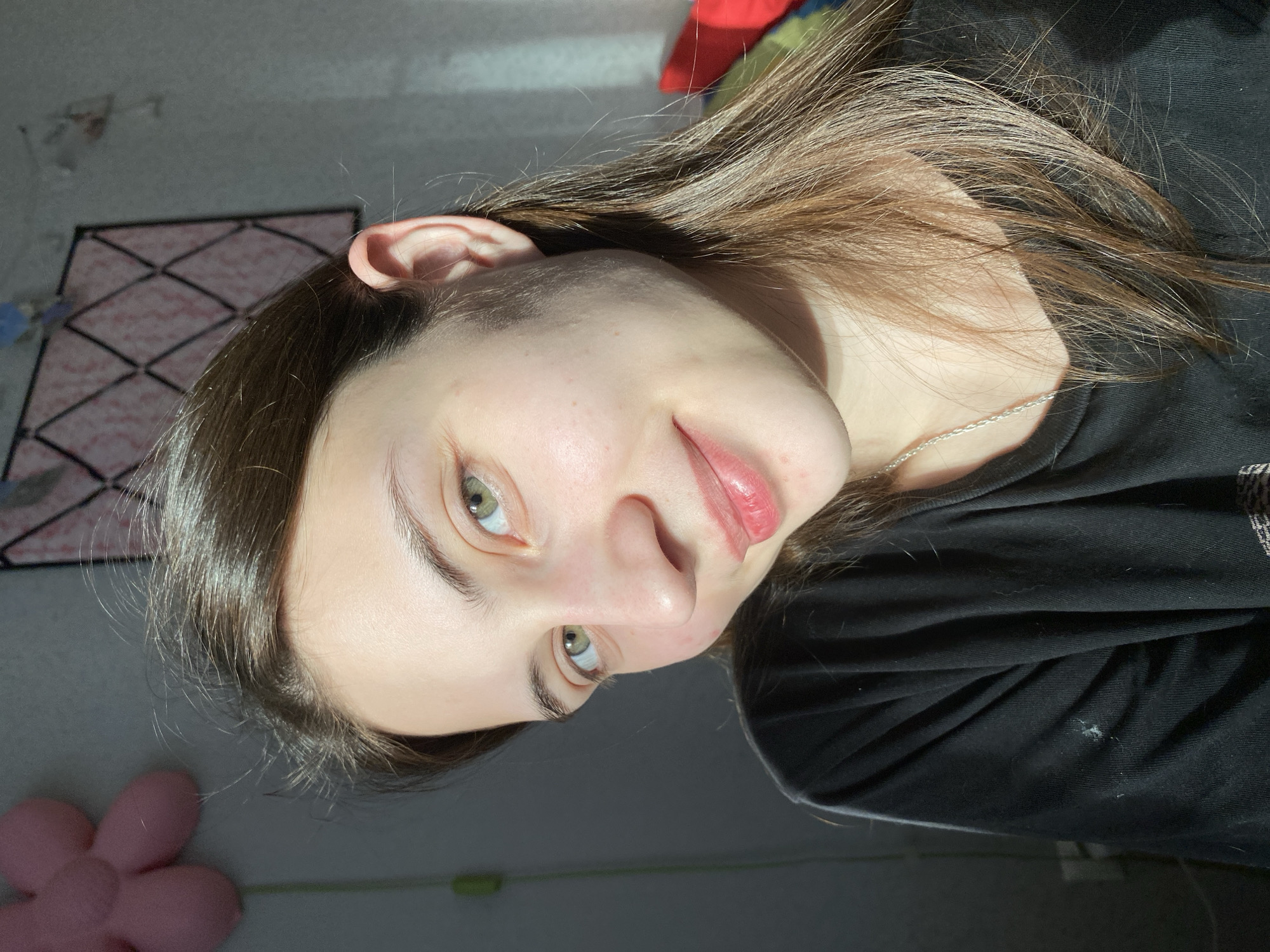 A young woman is shown in a close-up portrait with a slight tilt. She has long, straight brown hair, green eyes, and fair skin. She is wearing a black t-shirt and a silver chain around her neck. The background is a blurred combination of a light gray wall with a patterned object affixed, suggesting an indoor setting, possibly a room. A pink flower-shaped object is visible in the lower left corner. The lighting appears to be a combination of natural light and indoor lighting, with a bright, direct light source creating shadows on her face and clothing. There is no specific information or evidence to indicate a connection to the city of Votkinsk, Russia.