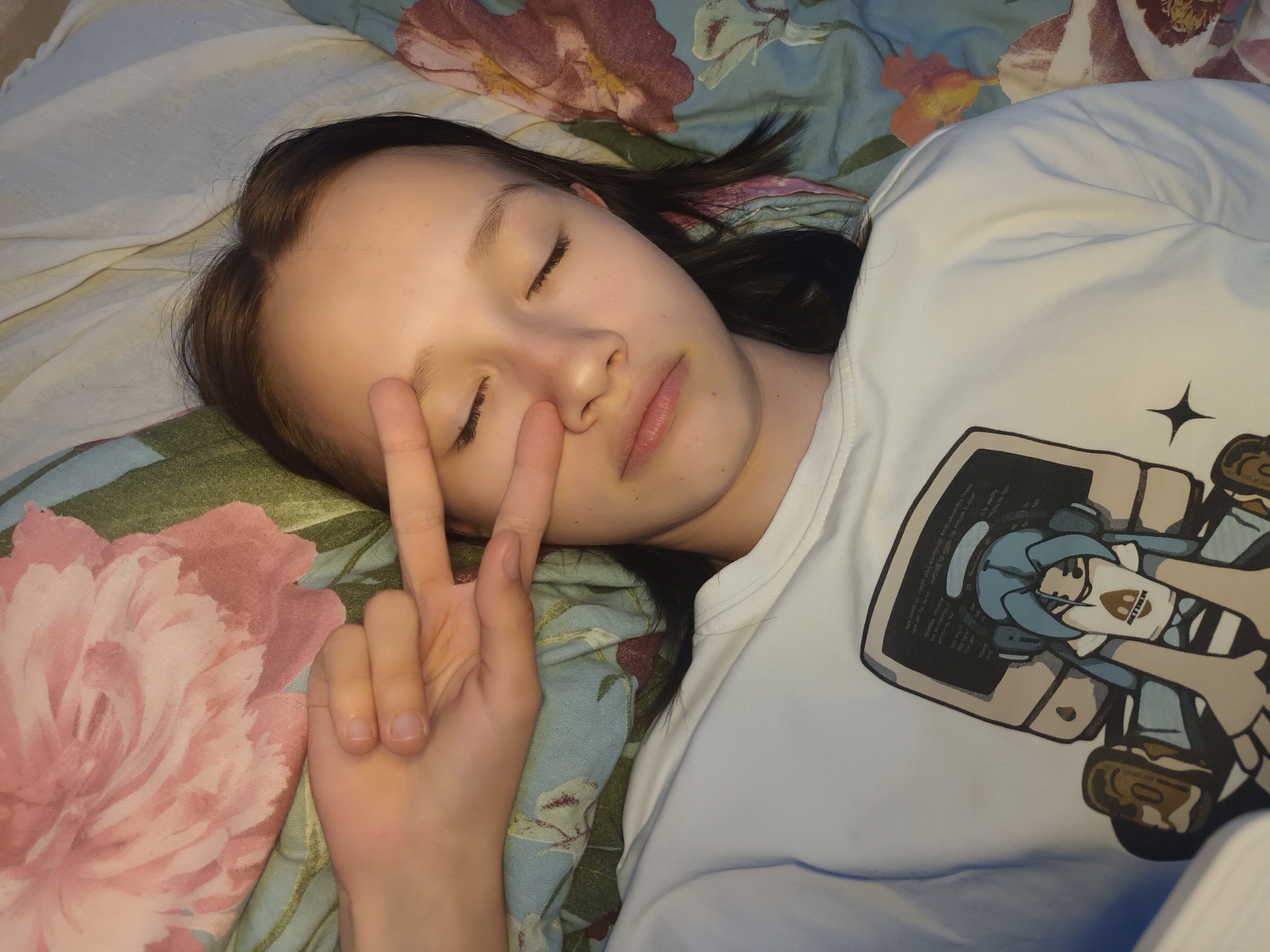 A young person, likely a female, is depicted lying on their side, seemingly asleep or resting peacefully, in an indoor setting in Ольховка, Russia. Their eyes are closed, and lips are slightly parted, conveying a serene expression. They have dark, straight hair spread across the pillow.
The person is wearing a light-colored (pale blue or off-white) short-sleeved T-shirt with a prominent graphic design on the chest. The graphic features an anime-style character with light blue hair, wearing a white shirt and a darker accessory, appearing to interact with or look at a monitor or screen. Small, illegible text is visible on the screen within the graphic, and a word that appears to be "GETHER" or "GET HER" is visible on a small patch on the character's clothing.
Their left hand is raised near their face, forming a peace sign (V-sign) with the index and middle fingers extended. The nails are short and unpolished. The background consists of a patterned pillowcase or duvet cover featuring large, soft pink peony-like flowers and green/blue foliage against a lighter background. The overall lighting suggests a subdued, possibly evening, indoor environment.