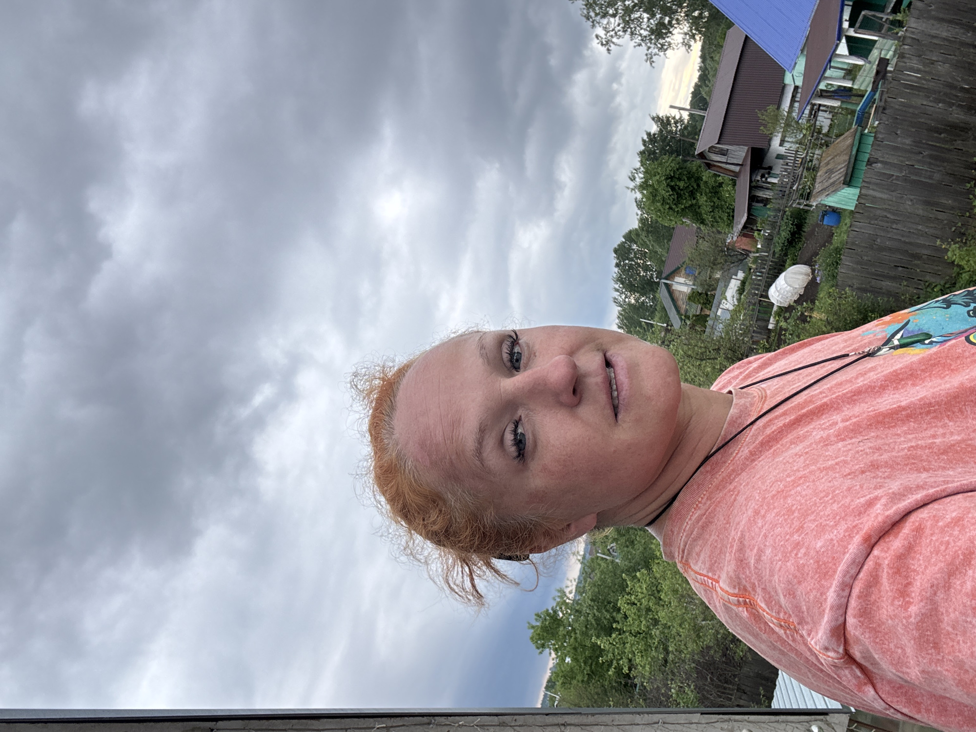 A selfie image shows a woman with red hair and blue eyes, wearing a pink t-shirt, with a necklace. She is outdoors, likely in a rural or suburban area. In the background are houses with different colored roofs, trees, and an overcast sky filled with gray clouds. The photograph was taken in an outdoor setting, specifically near a fenced yard or a building's edge. The angle suggests the photo was taken from a slightly elevated position. It is difficult to confirm the location is Chaikovsky, Russia, based solely on this image.