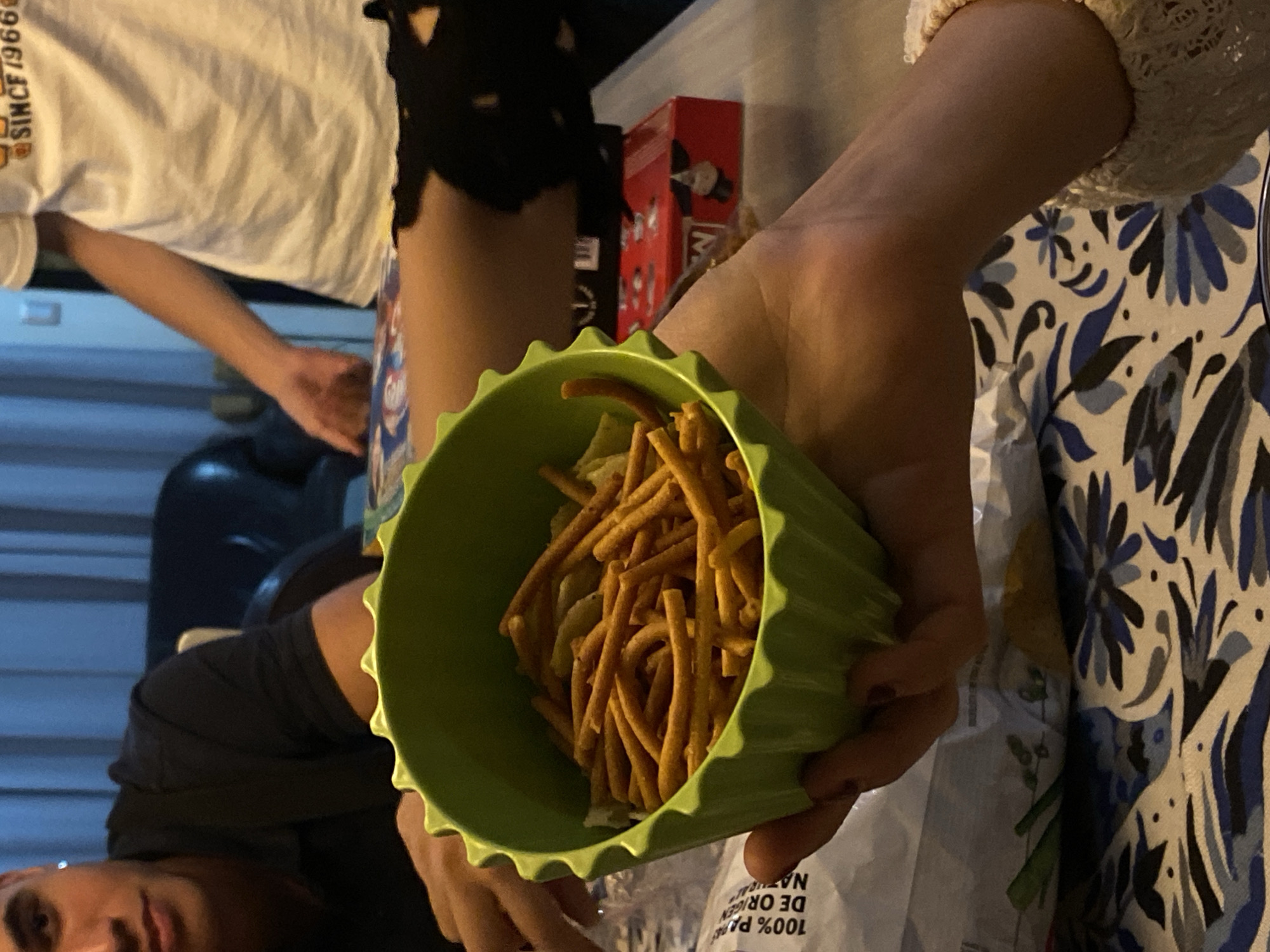 A high-angle shot depicts a person holding a green bowl filled with snacks, likely sticks, against a backdrop including parts of a white t-shirt with lettering, a person's legs and shorts, a red box, and a blue and white patterned surface. The photo's location is implied to be in Santa Clara Ocoyucan, Mexico, although direct evidence within the image to confirm the location is absent. Another person is partially visible in the bottom left corner.