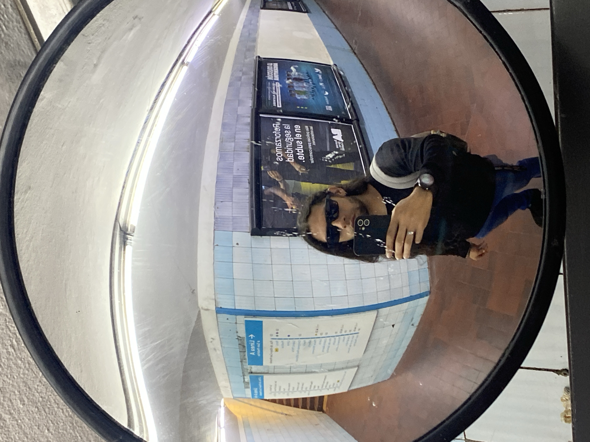 The image is a reflection in a convex mirror, showing a person taking a selfie in what appears to be a subway station in Buenos Aires, Argentina. The person is holding a smartphone with sunglasses reflecting in the screen. The reflection shows a tiled wall with posters, track, and lights. A watch on the person's wrist is visible. The ground is tiled.
