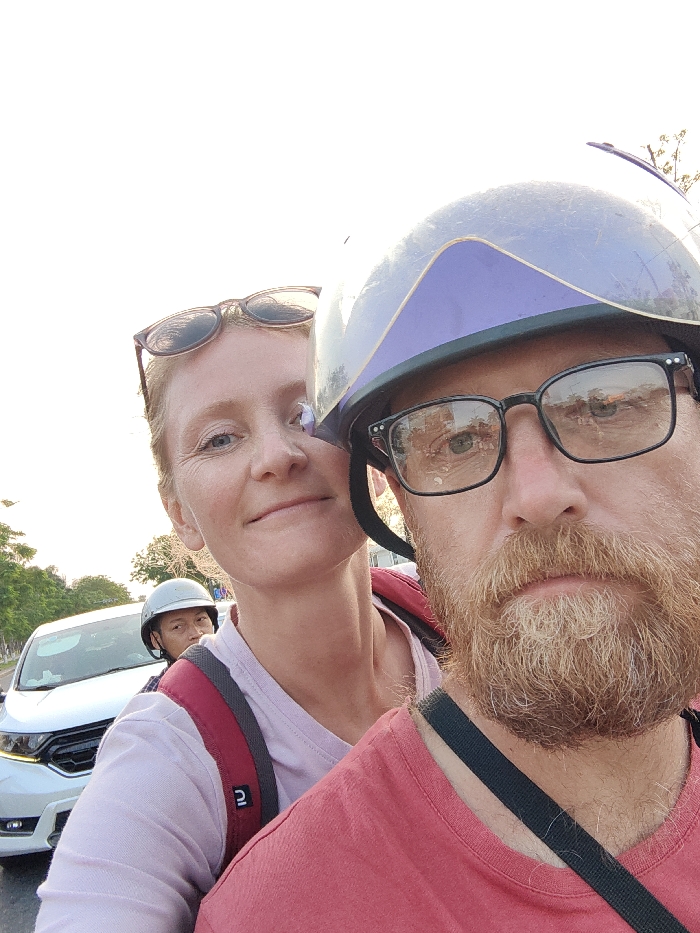 The scene depicts a close-up, daytime selfie taken on a street in Thuận Hóa Ward, Vietnam, showing two individuals on what appears to be a motorcycle or scooter, with other traffic in the background.
In the foreground, a Caucasian male with a reddish-brown beard and mustache is visible from the chin to the top of his head. He wears black-framed glasses, through which reflections of distant structures are visible, and a light grey helmet with a purple-tinted visor. He is dressed in a red t-shirt, and a black strap from an unseen object crosses his right shoulder. His gaze is directed towards the camera with a neutral expression.
Behind and to the left of the male, a Caucasian female is partially visible, showing her face, neck, and upper torso. She has light-colored hair, with sunglasses resting on her head, and displays a faint smile. She is wearing a light pink t-shirt and a red backpack with grey shoulder straps.
In the background, positioned behind the female, an Asian male wearing a light grey helmet is partially visible, appearing to be on another vehicle. To the far left in the background, the front portion of a white, modern-style car is discernible. The environment features a paved road, with sparse trees visible along the left side of the street under a bright, clear sky.