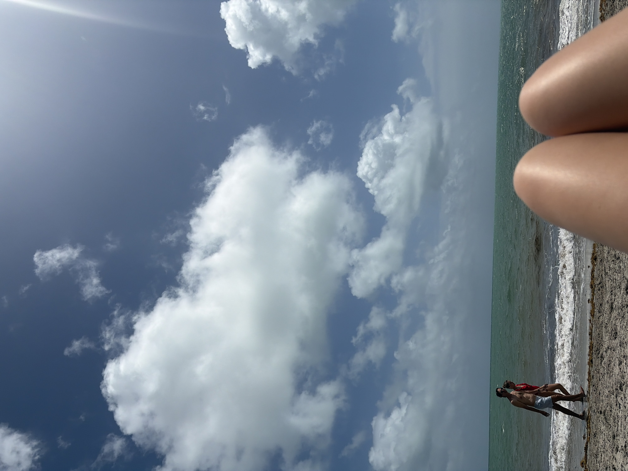 The image presents a scene at Key Biscayne, United States. A sunny sky with scattered white clouds fills the upper portion of the frame. The ocean horizon divides the mid-section, with waves breaking along the shoreline. Two individuals, one wearing red and the other blue swim suit, are walking on the beach. The lower right side of the frame features a set of legs.