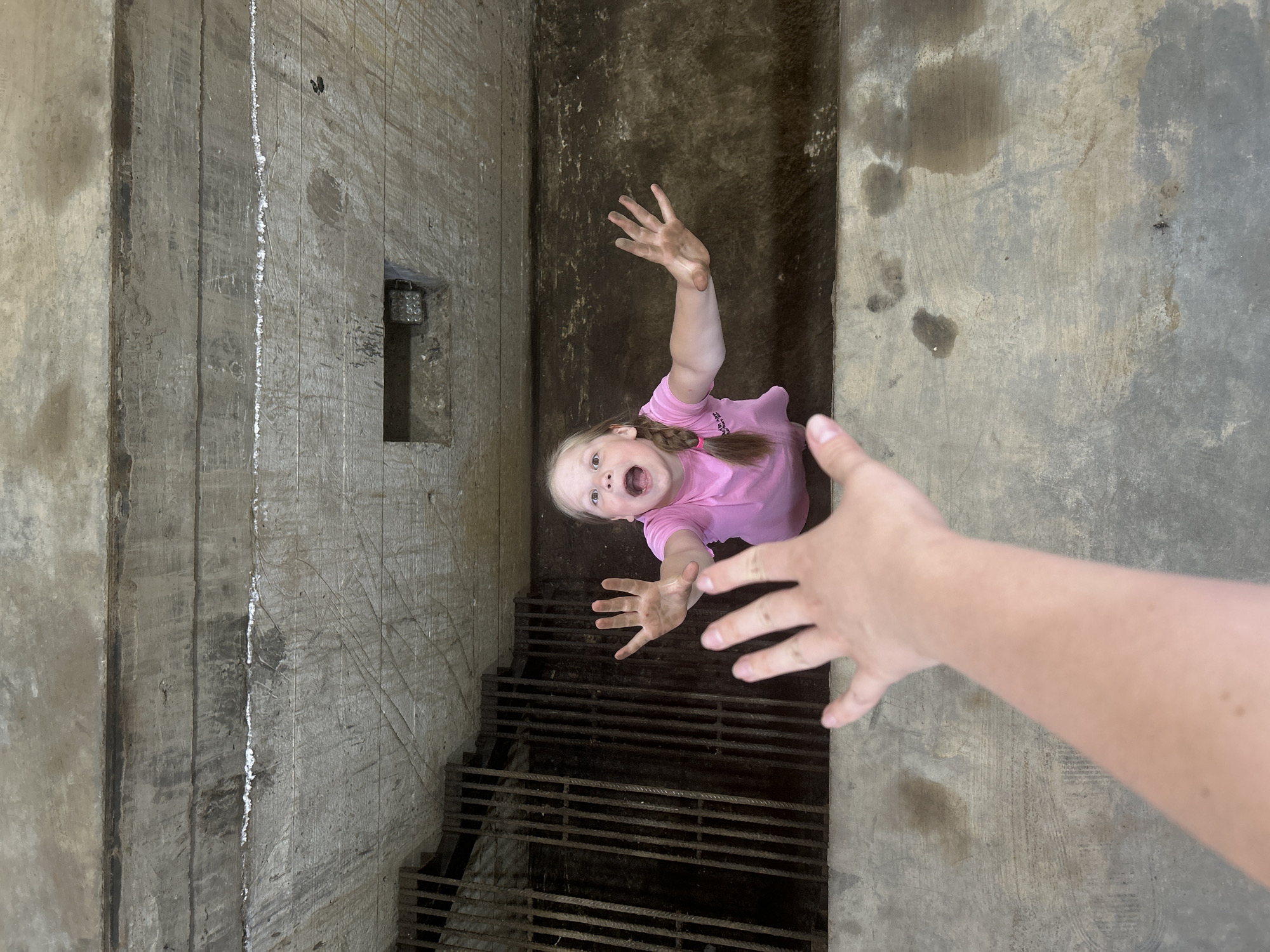 A low-angle, overhead shot depicts a child in a deep, concrete-walled pit or shaft. The child, wearing a pink shirt and with a braid in her hair, has both arms raised with hands open, reaching upwards. Her hands appear dirty. The child's face is visible, expressing an open-mouthed shout. A blurry human hand reaches down towards the child from above. A grid-like metal structure forms the floor of the pit, suggesting a drainage or access point. The pit's walls are rough, textured concrete, likely weathered or aged. The location is indicated as Chaikovsky, Russia.