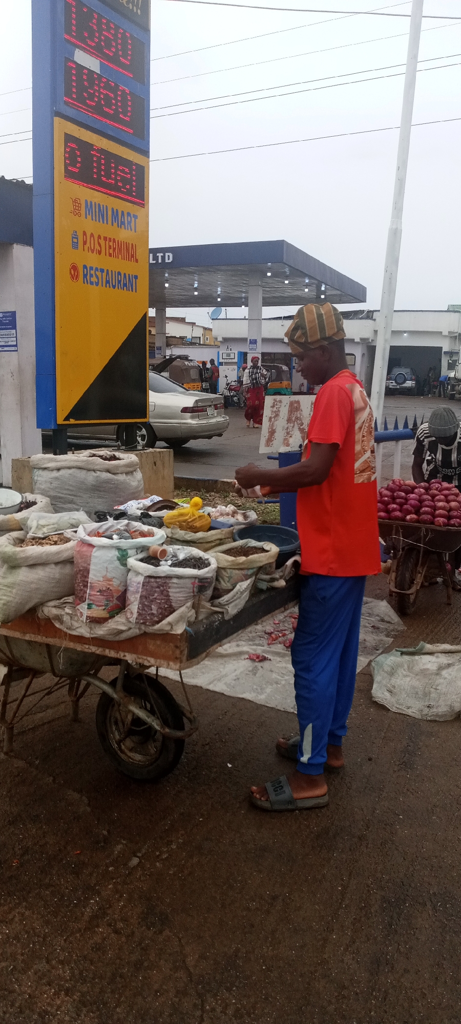 A man is selling goods from a cart in front of a petrol station in Jos, Nigeria. The petrol station has a blue and yellow sign with prices listed. The cart is loaded with sacks of various items. The man is wearing a red shirt, blue pants, sandals, and a striped cap. He appears to be sorting through items. Another cart filled with red onions is nearby, and the scene is on a paved road in an urban environment. In the background, there's the fuel station and cars. The sky is overcast.