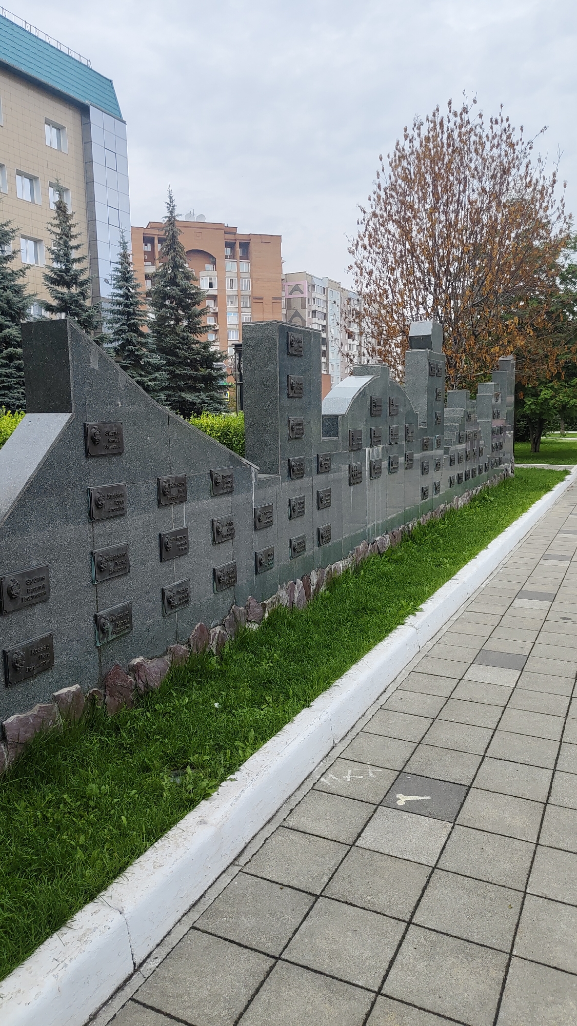 The image shows a memorial with plaques on a granite structure that resembles a city skyline. The memorial is situated in a park-like setting with green grass, trees, and buildings in the background. The plaques appear to be inscribed with names and possibly dates, suggesting it is a memorial to individuals. The foreground features a paved walkway with square concrete tiles. The overall scene conveys a somber and reflective atmosphere. This memorial is located in the city of Krasnoyarsk, Russia.