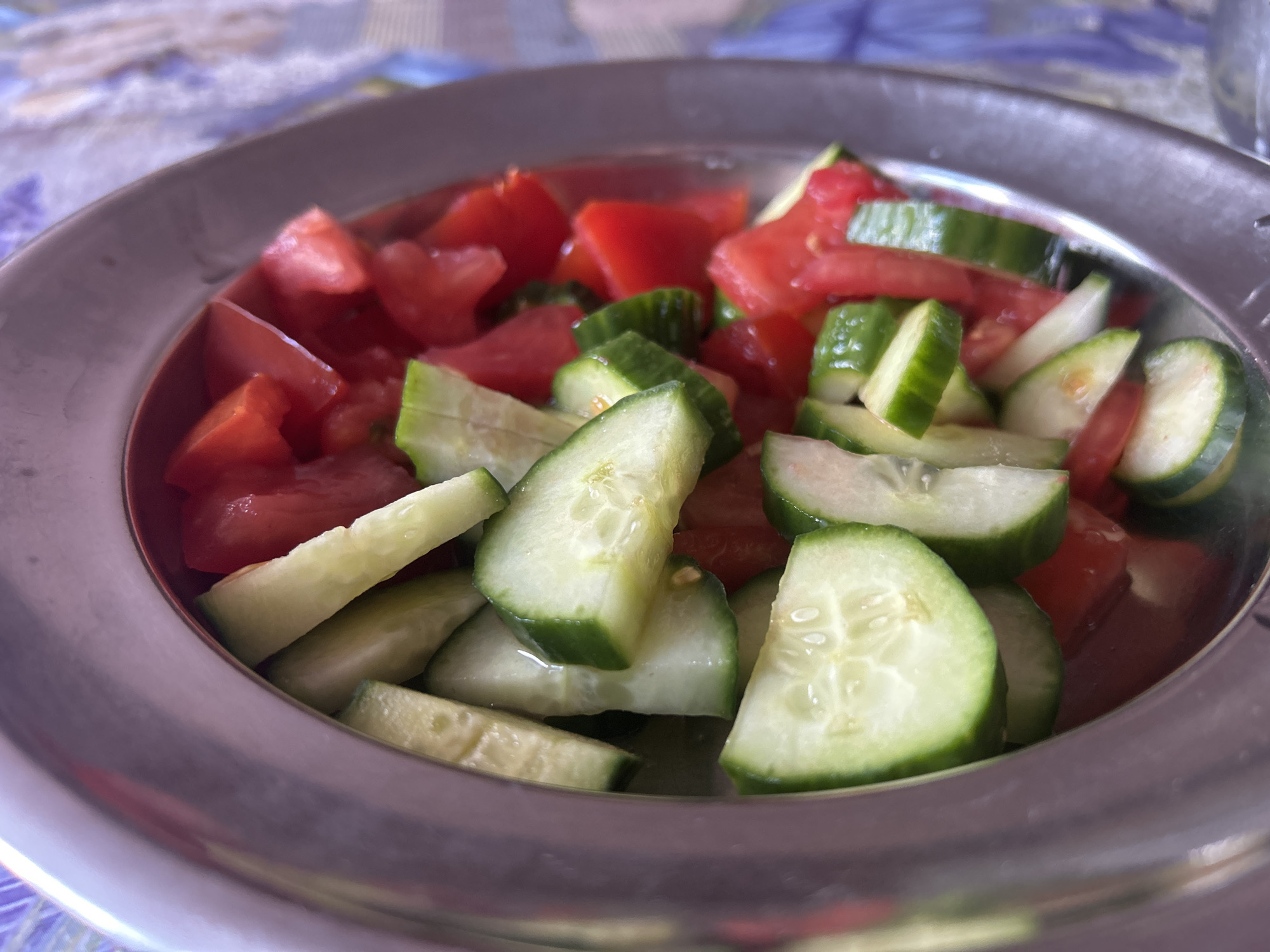 A close-up shot depicts a metal bowl filled with a salad of diced tomatoes and cucumber slices. The bowl, with its slightly curved lip, sits on a surface with a patterned tablecloth, potentially in a home or restaurant setting. The salad consists of bright red tomato pieces and green cucumber slices, appearing fresh and moist. Given the request to include the city of Dve Mogili, Bulgaria, one could infer that this salad is being served or consumed in that location. There are no people in the frame.