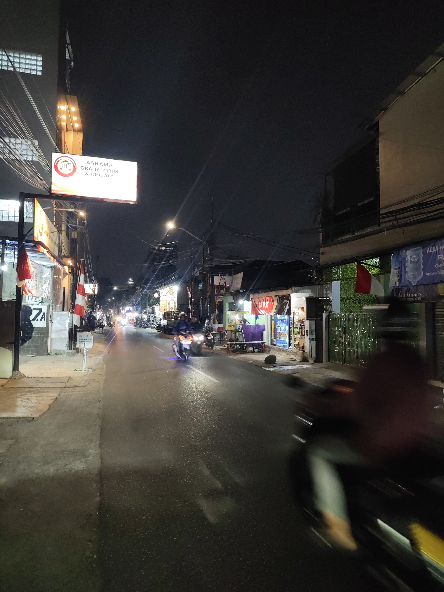 The image captures a street scene at night in South Jakarta, Indonesia. A brightly lit billboard for "ASRAMA GRAHA YATIM & DHU'A" is prominently displayed on the left side of the street. The road is wet, suggesting recent rain or a damp environment. Several motorcycles are visible, with one in the foreground blurred due to motion. Other motorcycles and a few pedestrians are present further down the street, illuminated by streetlights and shop signs. The buildings lining the street appear to be a mix of commercial establishments and residences, with electrical wires forming a dense network overhead. The overall atmosphere is that of a busy, urban night.