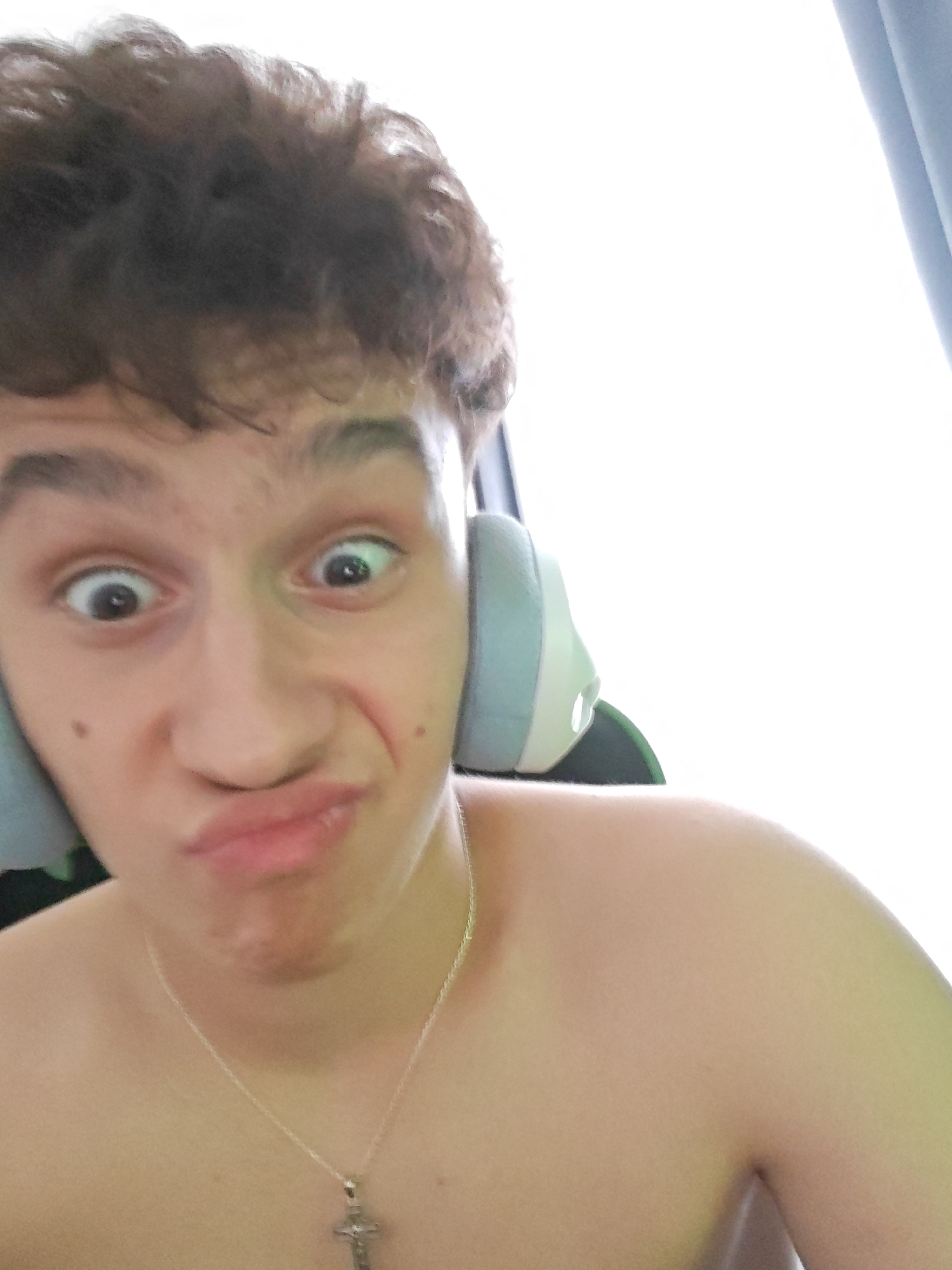 A young person with curly brown hair makes a goofy face with wide eyes and pursed lips while wearing headphones. The person is shirtless, wearing a silver chain with a cross pendant. The background is mostly out of focus, with a bright white area and a hint of a blue curtain. The image appears to be a selfie taken indoors.