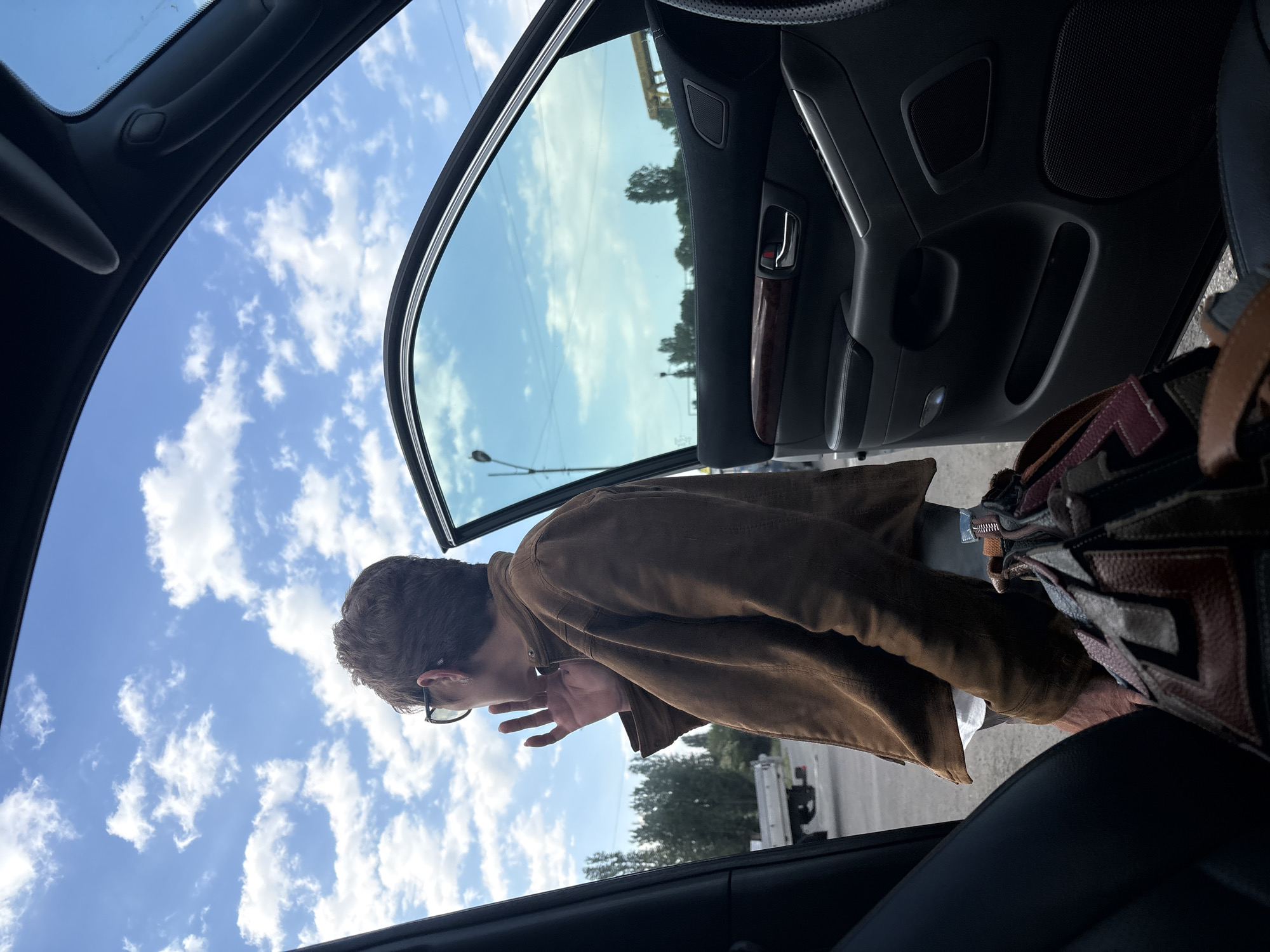 A medium shot taken from inside a vehicle, likely a car, angled towards the open driver-side door. Outside, a person wearing glasses and a brown jacket leans slightly, seemingly looking into the car. The sky is visible through the open door, filled with white clouds. A bag is partially visible near the person. The interior of the car is visible, including the door panel and the steering wheel. The location cannot be determined to be Burgas, Bulgaria from the visual information alone.
