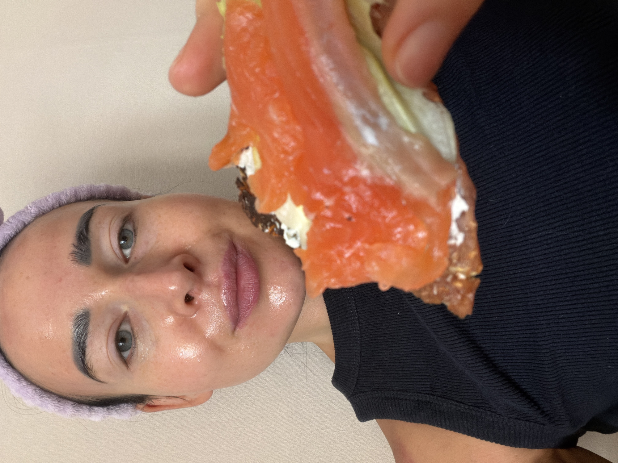A fair-skinned woman is holding a sandwich with salmon in front of her face. She is wearing a purple headband and a black top. Her face appears shiny, perhaps due to skincare products or sweat. The sandwich appears to be open-faced and contains dark bread, a white spread (possibly cream cheese), and salmon. The background is a plain, light-colored wall. It is not possible to determine the location in the city of Saint Petersburg, Russia based on the provided image.