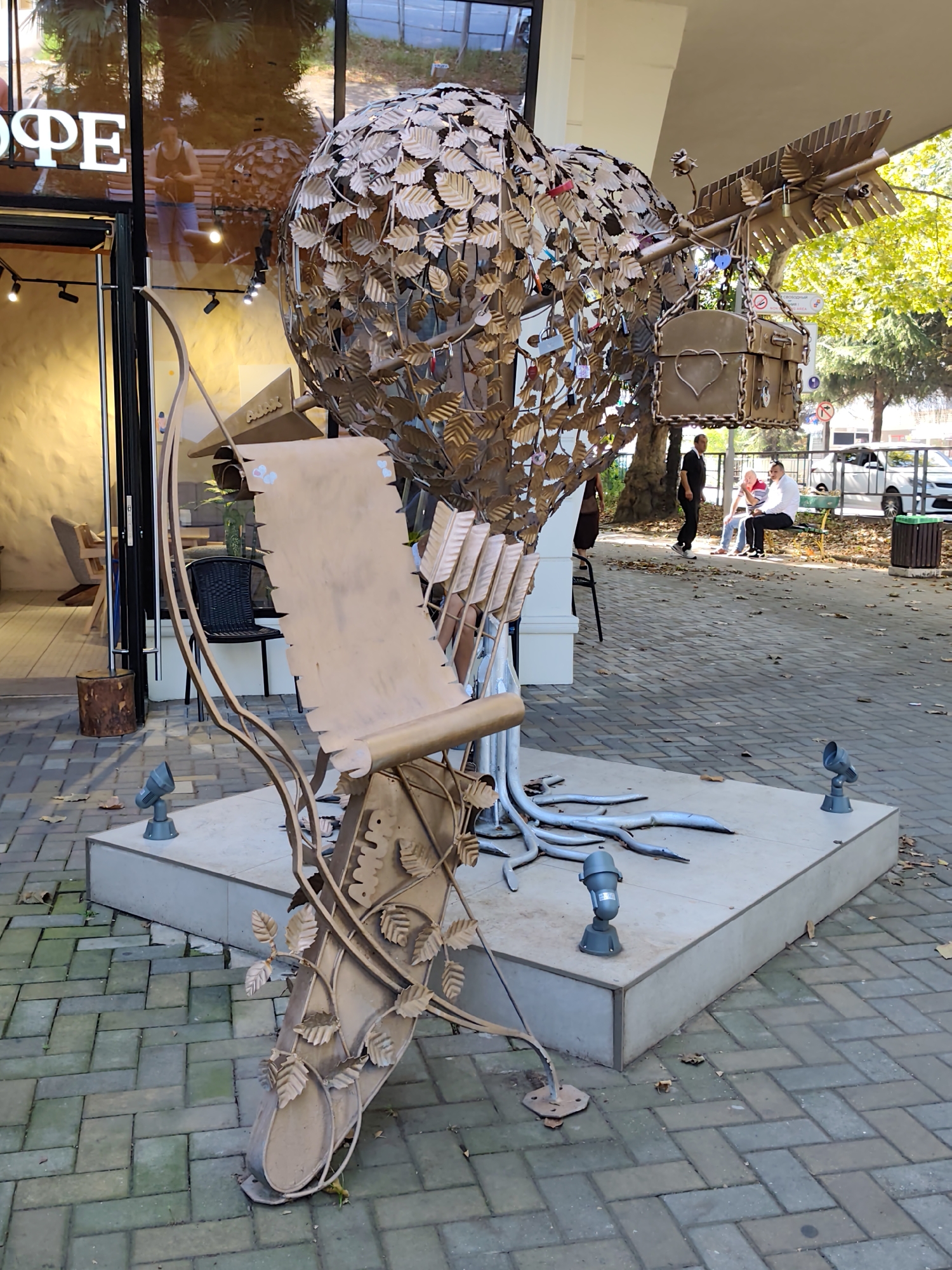 A metallic tree sculpture with leaf-shaped ornaments and a scroll-like element is positioned on a paved walkway outside a cafe with a "COFE" sign. Several padlocks are attached to the sculpture's branches. In the background, three men are sitting on a bench, with a white car and a green trash bin visible further down the street. The scene appears to be in Sochi, Russia, based on the context provided.