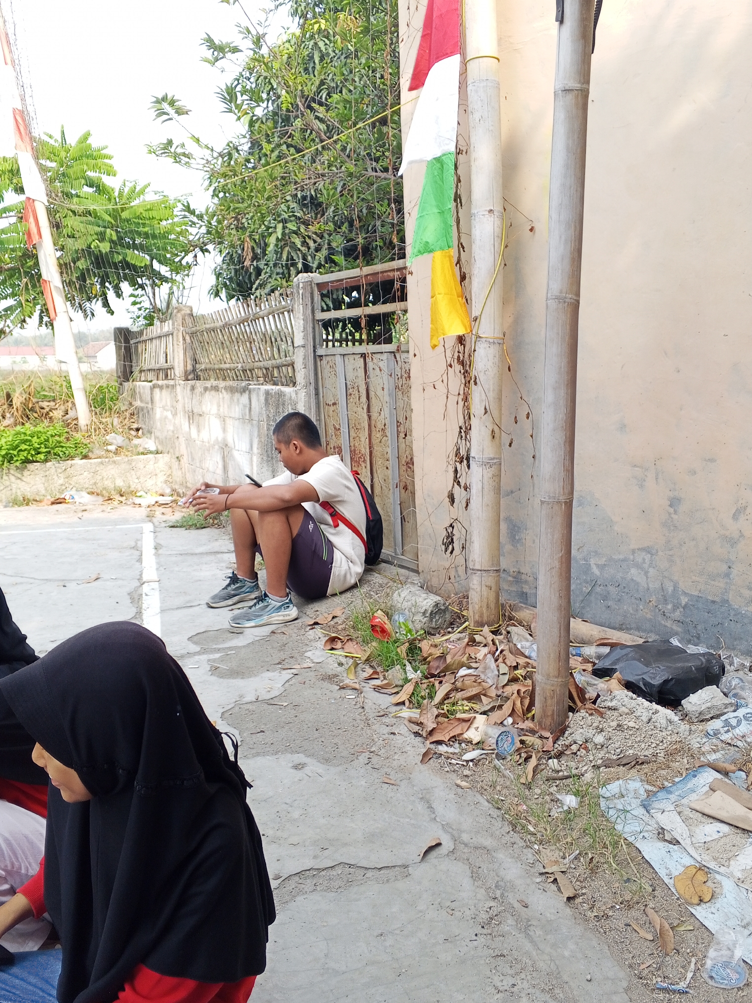 A medium shot captures a young man sitting on a concrete sidewalk, looking down at his phone. He is wearing a white t-shirt, dark shorts, and sneakers, with a black backpack slung over his shoulders. To his left, partially visible in the foreground, is a person wearing a black hijab and a red shirt. Behind the young man, a weathered metal gate leads to a yard with a bamboo fence and lush green trees. A tall bamboo pole stands to the right of the gate, adorned with red, white, green, and yellow flags. The ground is littered with dry leaves, trash, and debris, including plastic bottles and a black garbage bag. The scene appears to be outdoors, possibly in a residential area or near a public space in Ciruas, Indonesia. The overall impression is one of casual waiting or resting in a somewhat unkempt environment.