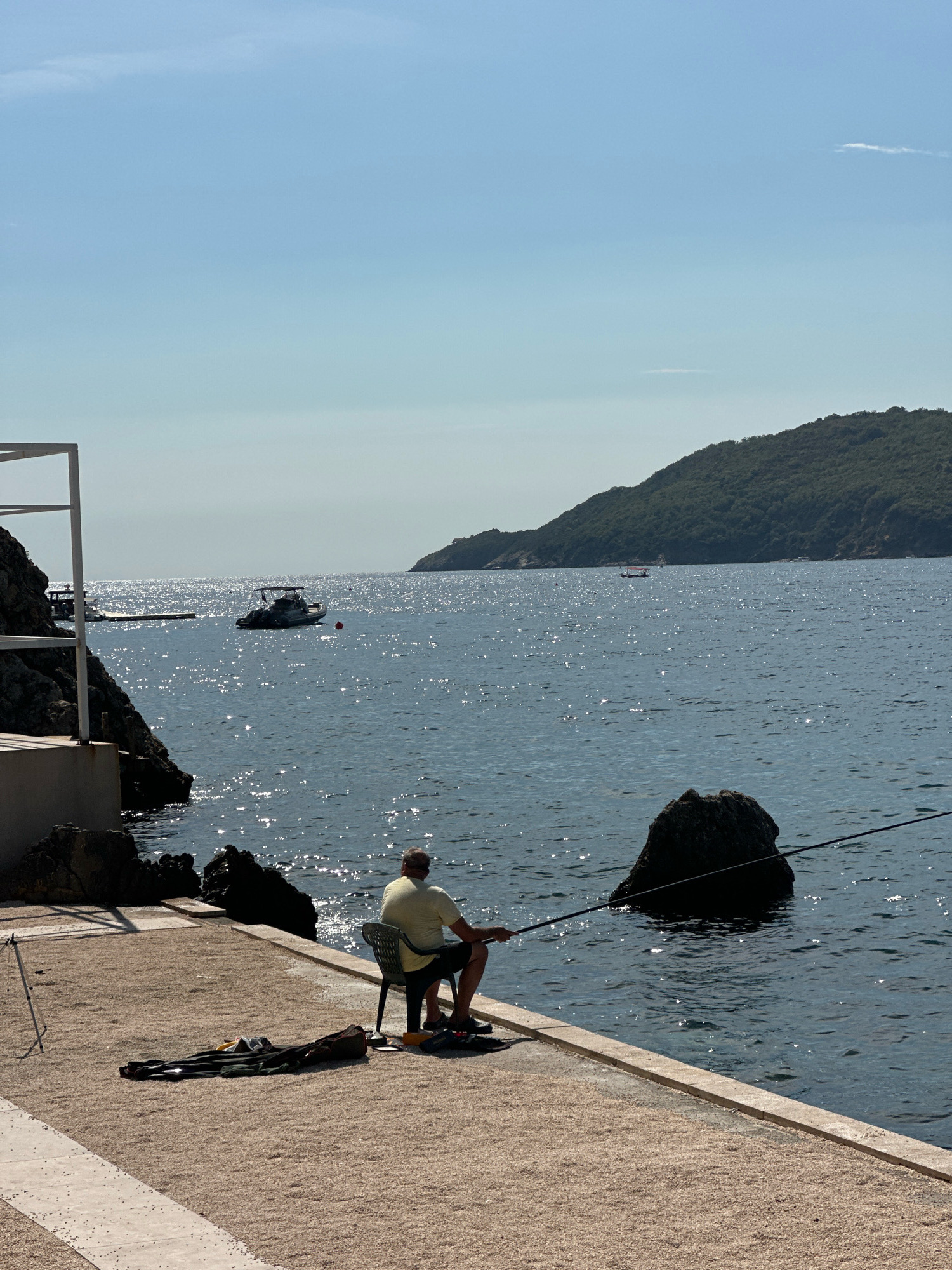 The image depicts a man fishing from a paved waterfront walkway in Budva, Montenegro. The man is seated in a dark chair, wearing a light yellow t-shirt and dark shorts, holding a fishing rod that extends into the calm, sparkling blue sea. In the foreground, a fishing bag and other equipment lie on the ground next to a tripod. The walkway is made of light-colored gravel with white concrete borders. To the left, a white structure with vertical beams suggests part of a modern building or canopy. Rocky outcroppings are visible along the water's edge. In the middle distance, a motorboat is anchored, and a red buoy floats nearby. Further out in the sea, a distant green, tree-covered island with a hilly terrain dominates the background. The sky is a clear, pale blue, with faint wisps of clouds visible near the horizon. The scene is bathed in bright sunlight, creating reflections on the water's surface.