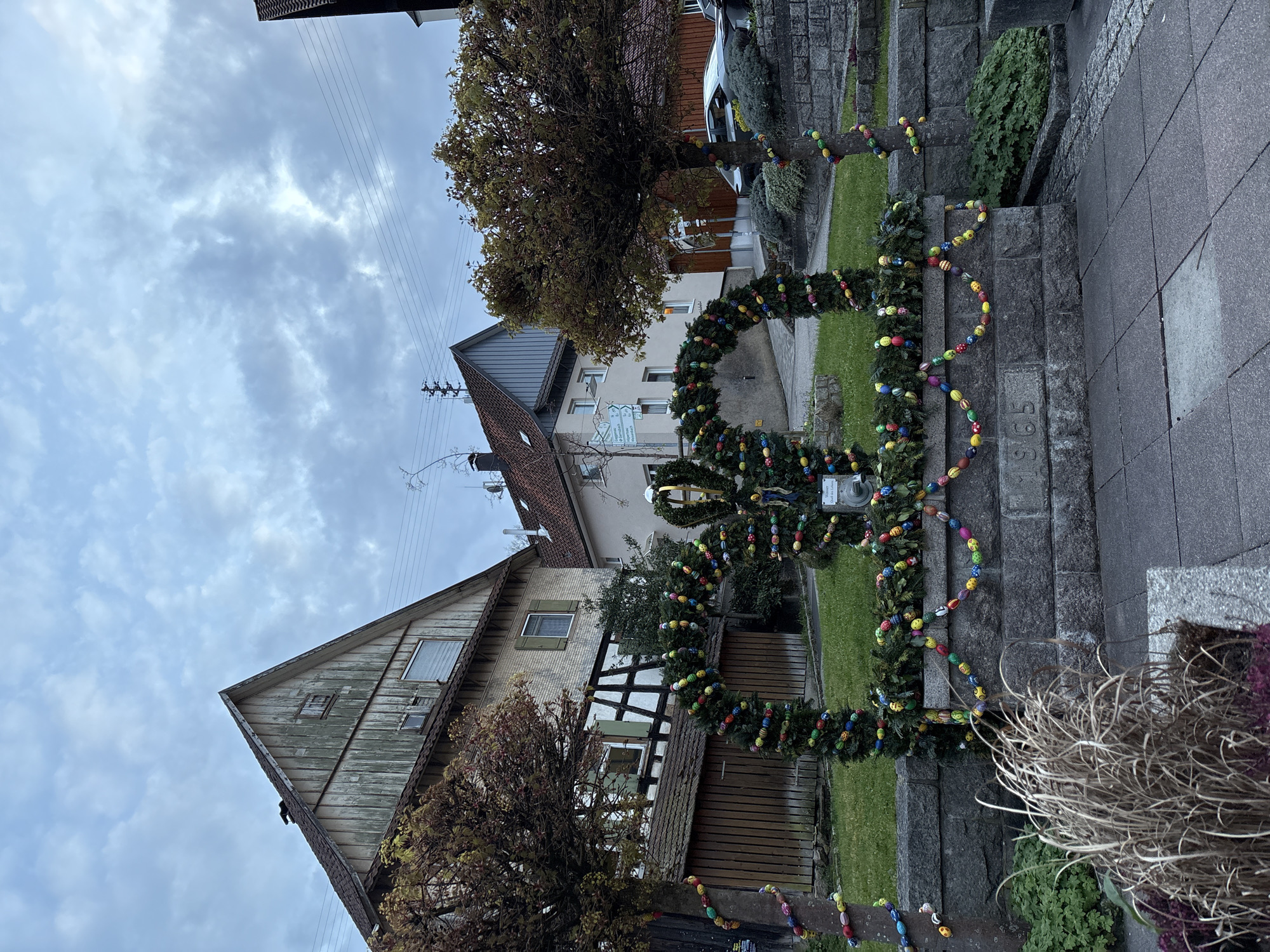 The image shows a street scene in Forbach, Germany, with a stone staircase decorated for Easter. The staircase is adorned with green garlands embellished with colorful Easter eggs. The garlands are arranged in a series of arches and loops, creating a festive display. In the background, there are buildings of various architectural styles, including a traditional timber-framed house and a more modern structure. Bare trees are visible throughout the scene, and power lines run overhead. The sky is overcast, suggesting a cloudy day.