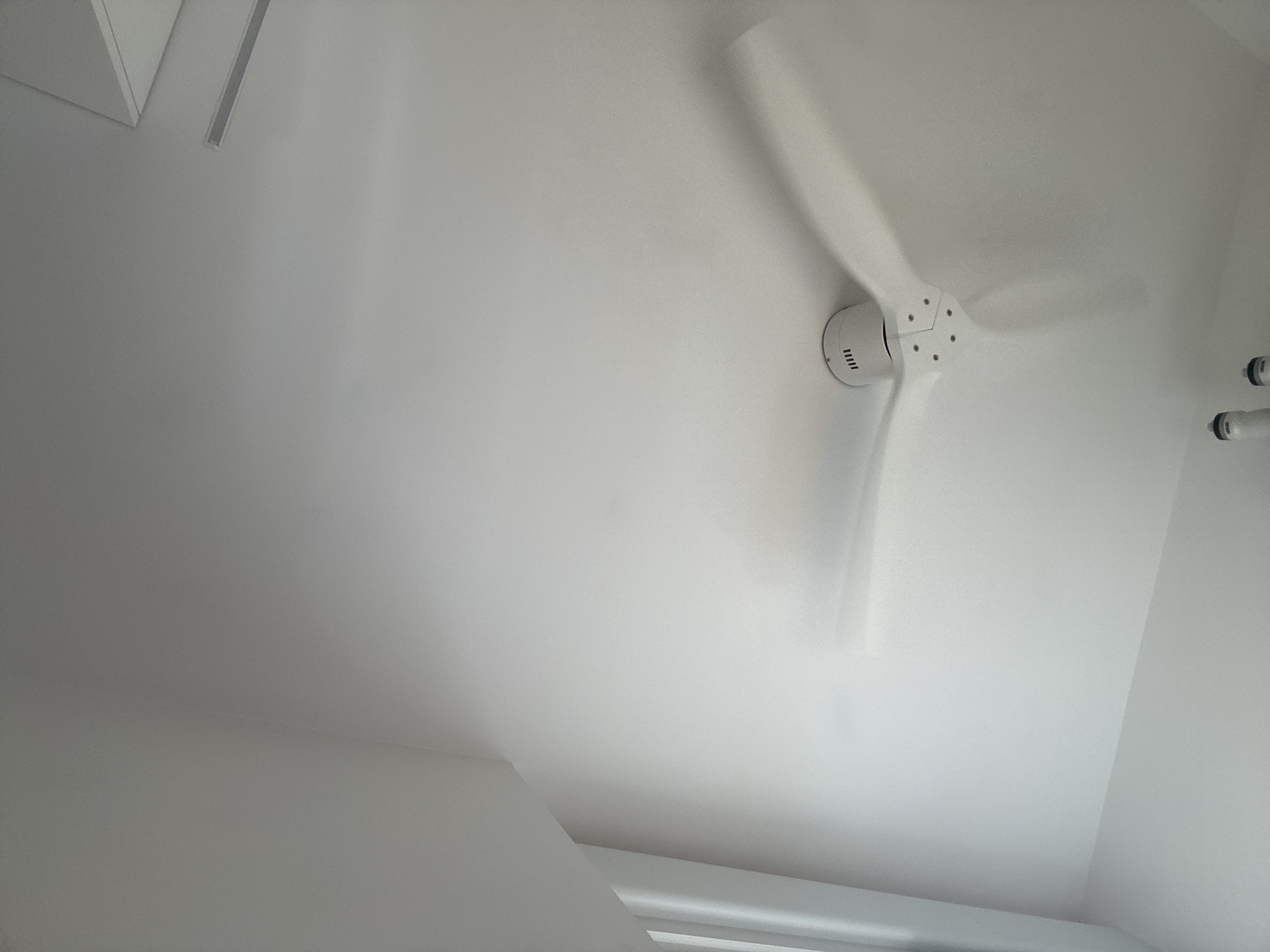 The image depicts the interior of a modern room with white walls and a white ceiling, creating a minimalist aesthetic. A white ceiling fan with three blades is prominently featured, positioned on the left side of the frame and angled towards the viewer. The fan appears to be in motion, as evidenced by the slight blur of the blades. Two cylindrical objects, possibly lights or sensors, are visible on the far left edge of the image. The lighting suggests an indoor environment, with light sources originating from above and to the right, casting soft shadows and highlighting the textured surface of the walls. In the upper right quadrant, a section of the ceiling appears to be angled, suggesting an architectural feature such as a sloped ceiling or a skylight. The overall impression is one of clean lines and a bright, airy space. There is no indication of the specific location within the city of Murcia, Spain.