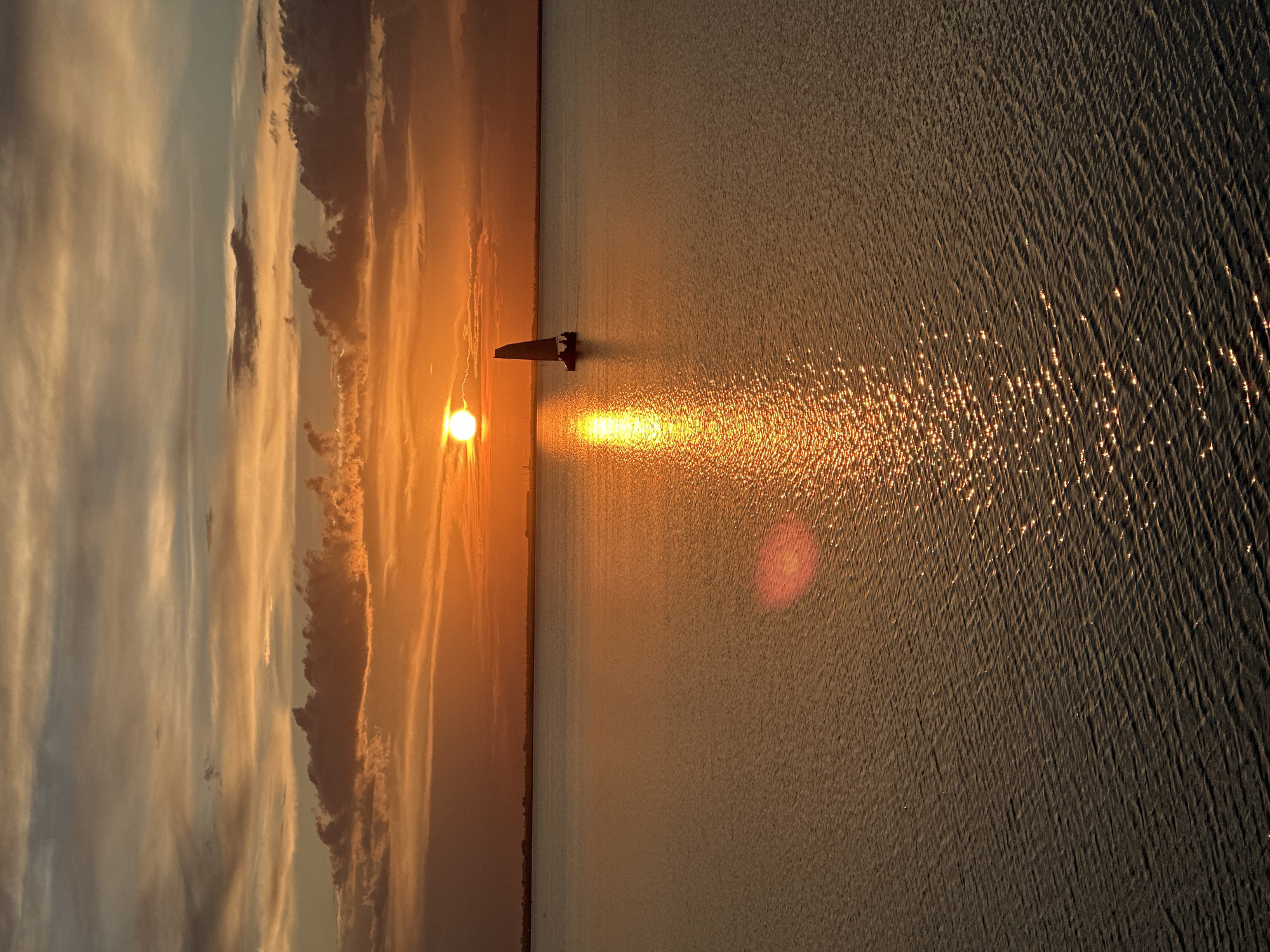 A sunset over a body of water in Kokshetau, Kazakhstan, features a sailboat with two figures on board. The sun, a bright orb, is low on the horizon, casting a warm, orange glow across the sky and water. Clouds are scattered across the sky, tinged with orange and yellow hues. The water is calm, with gentle ripples reflecting the light of the setting sun. A faint shoreline is visible in the distance.