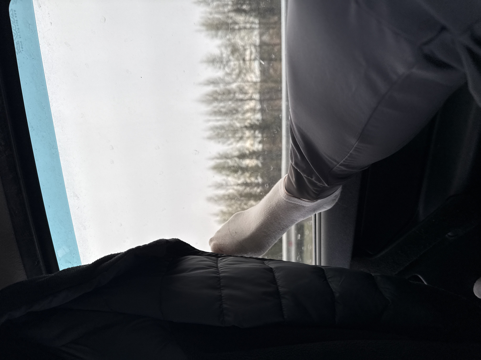 The image depicts a person's foot, clad in a white sock, resting on the interior window ledge of a vehicle, likely during travel. The leg, partially covered by light gray fabric, is positioned in a relaxed manner. In the foreground, a dark, quilted jacket or blanket is visible, suggesting comfort or protection from cold.
Through the window, a bright, overcast sky dominates the view, with faint water droplets or condensation visible on the glass surface. Below the sky, a dense line of blurred conifer trees indicates a wooded landscape, possibly seen from a moving vehicle. The diffuse lighting suggests an overcast day, without direct sunlight.
The overall scene conveys a sense of quiet travel and repose. Faint, mirrored text, likely a manufacturer's mark or safety standard, can be seen on the upper left corner of the window, including characters such as "AAA BUH" and "01.110-V03.V02". Given the location context of Pokrovskoye, Russia, the coniferous trees and overcast weather align with the region's climate.