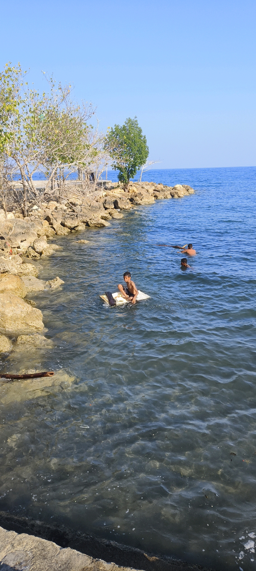 The image depicts a sunny day at the waterfront in Waisai, Indonesia. In the foreground, a young boy is seated on a makeshift raft made of white foam board and dark wood, floating in clear, shallow water. He is wearing dark shorts and is looking towards the right side of the frame. Further out in the water, two other individuals are visible swimming. One is partially submerged, with only their head and shoulders showing, and is holding onto a dark object that appears to be a long piece of wood or debris. The other swimmer's head is also visible above the water. The shoreline is lined with large, light-colored rocks and sparse vegetation, including trees with sparse foliage. The background shows a calm, blue ocean extending to the horizon under a clear blue sky. The overall scene suggests a recreational activity on a warm day by the sea.