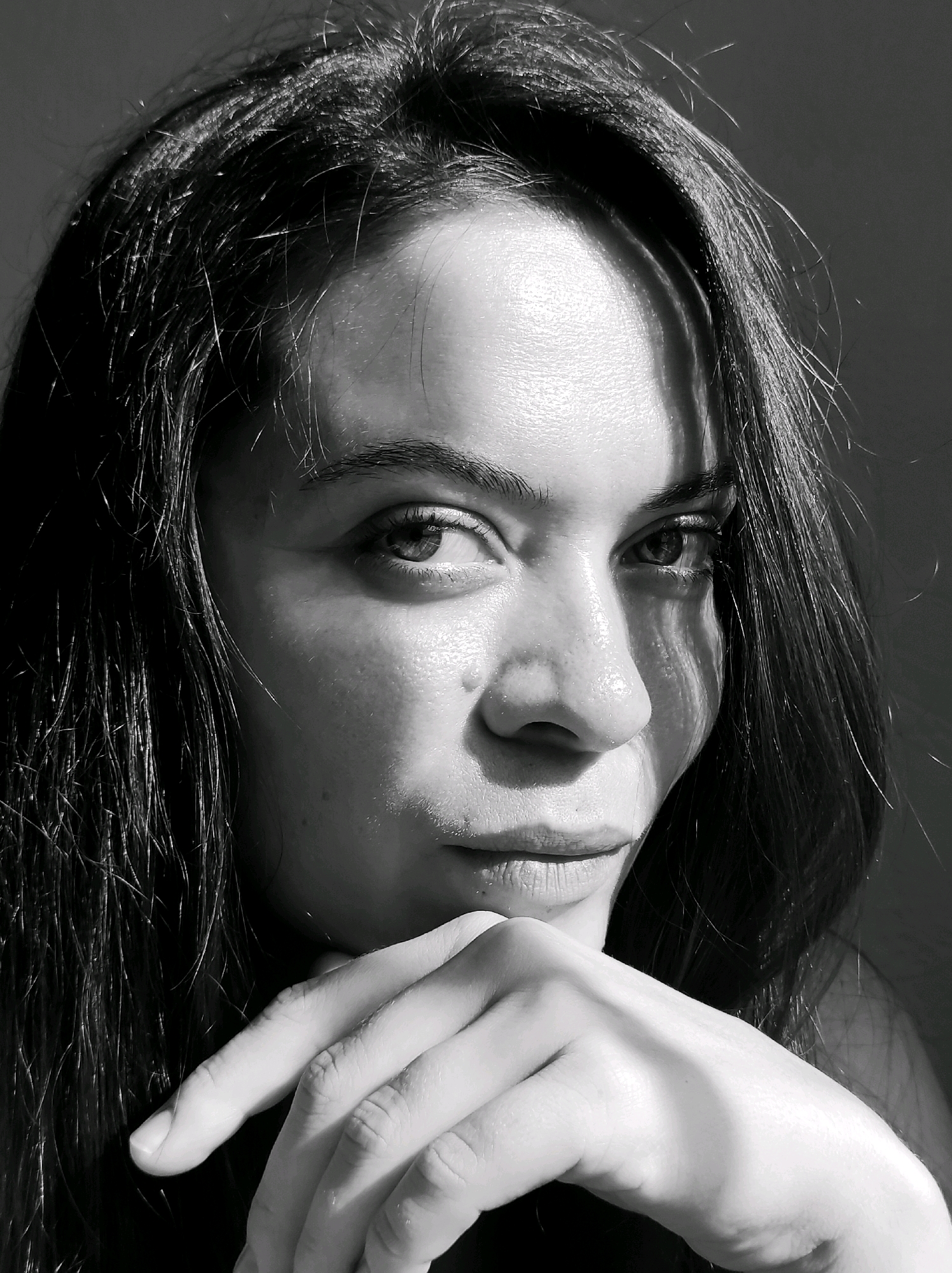 A black and white, close-up, waist-up photograph shows a woman with long, dark hair. The woman is leaning her chin on her hand, with her fingers gently touching her chin and mouth area. Her gaze is directed slightly upwards and to the viewer's left. Dramatic shadows fall across her face, highlighting her features and creating a moody atmosphere. The background is dark and indistinct. The woman's skin appears smooth with visible pores. There is a light reflection on her forehead and cheek. This image is not taken in Chaikovsky, Russia.