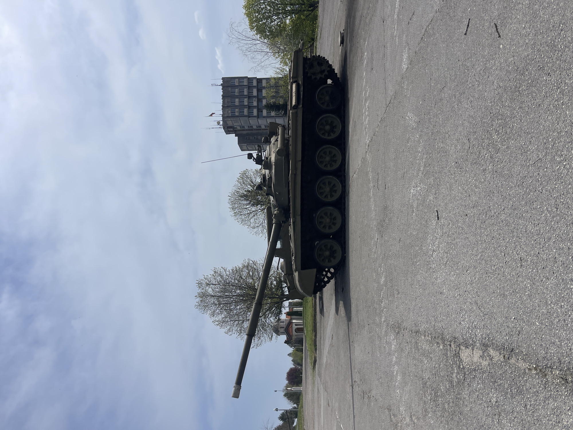 A medium-high angle shot, rotated 90 degrees counter-clockwise, shows a green T-72 main battle tank parked on a gray asphalt surface. Behind the tank, a tall, multi-story concrete building is visible, and to the right of the building are green trees and a light blue sky with scattered clouds. Other buildings are partially obscured by trees to the left of the tank.
Based on the architecture of the building in the background and the presence of a tank, this image is very likely taken in Veliko Tarnovo, Bulgaria at the Tank Museum. The tank is likely part of the open-air exhibit there.