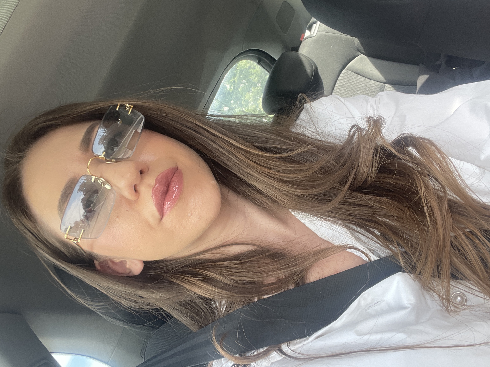 The image is a selfie taken inside a car. A woman with long brown hair is wearing sunglasses and a white shirt. She is wearing a seatbelt. The car appears to be moving, as can be deduced from looking at the trees through the window. The location cannot be determined to be Sofia, Bulgaria, from the image alone.