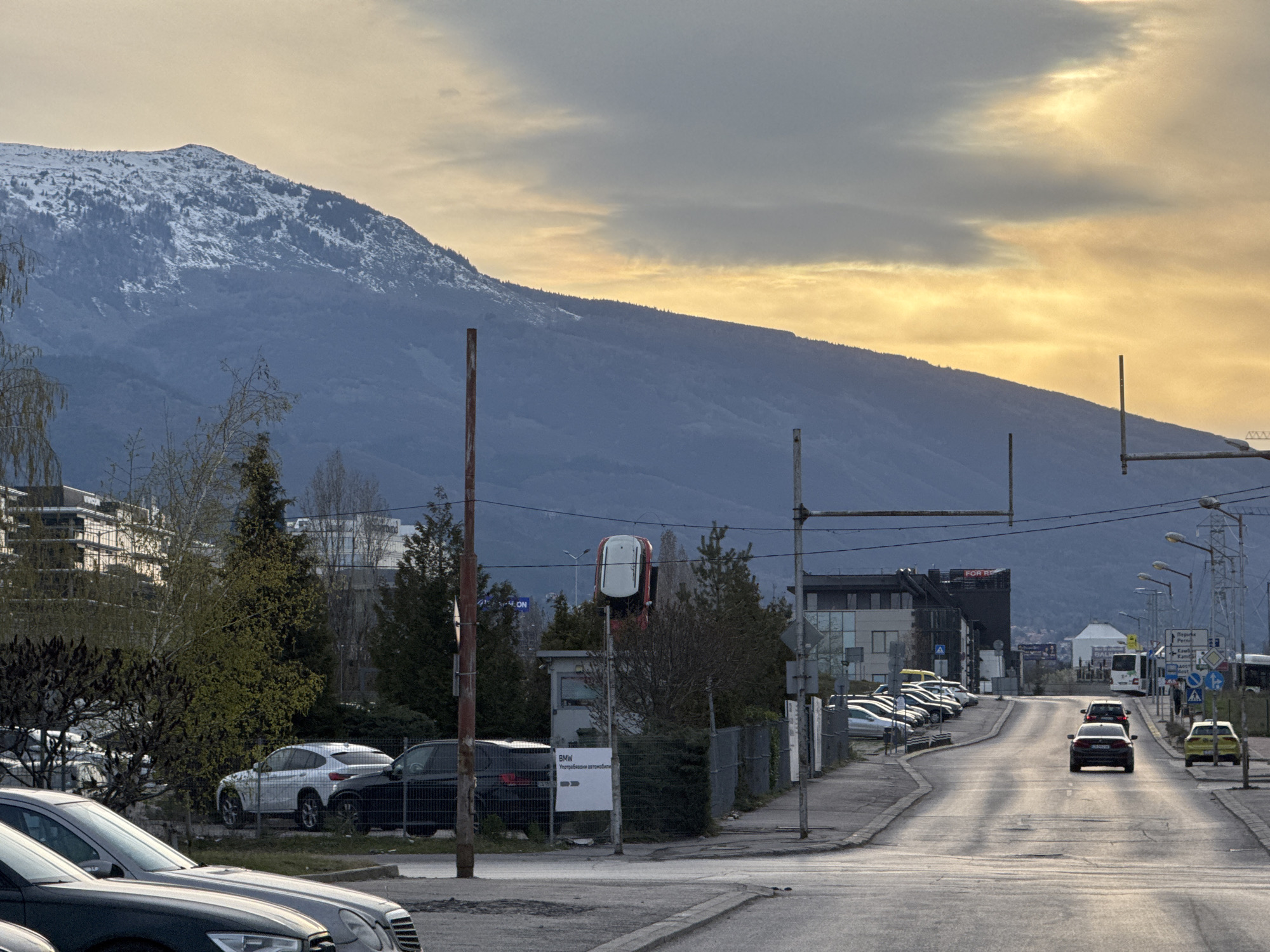 The image depicts a street in Sofia, Bulgaria, facing toward Vitosha Mountain. The road slopes gently uphill, with cars parked on the left side and traffic moving in both directions. Several cars are visible on the road, including a red car and a yellow car. On the left side of the road, there are several commercial buildings and a car dealership with a sign indicating "BMW". Overhead power lines stretch across the road, supported by metal poles. The sky is overcast with a yellow hue near the horizon, suggesting dawn or dusk. Vitosha Mountain dominates the background, with patches of snow visible on its peak.