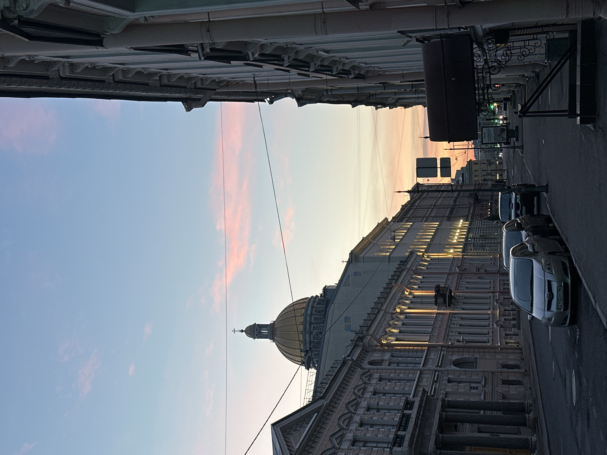 This is a vertical, eye-level, outdoor, daylight shot of a city street at dusk. The sky is a pale blue transitioning to pinkish-orange near the horizon. In the distance, a large, golden-domed building, likely a cathedral or church, is visible, topped with a cross. The street is lined with grand, ornate buildings with classical architecture. The buildings on the left feature columns and decorative detailing, while the building on the right is more minimalist in its ornamentation but still grand. The windows of the buildings are illuminated, suggesting interior lights are on. Several cars are parked along the street, including a silver hatchback in the foreground with a license plate visible. Power lines crisscross the sky. The overall atmosphere is serene and historic.