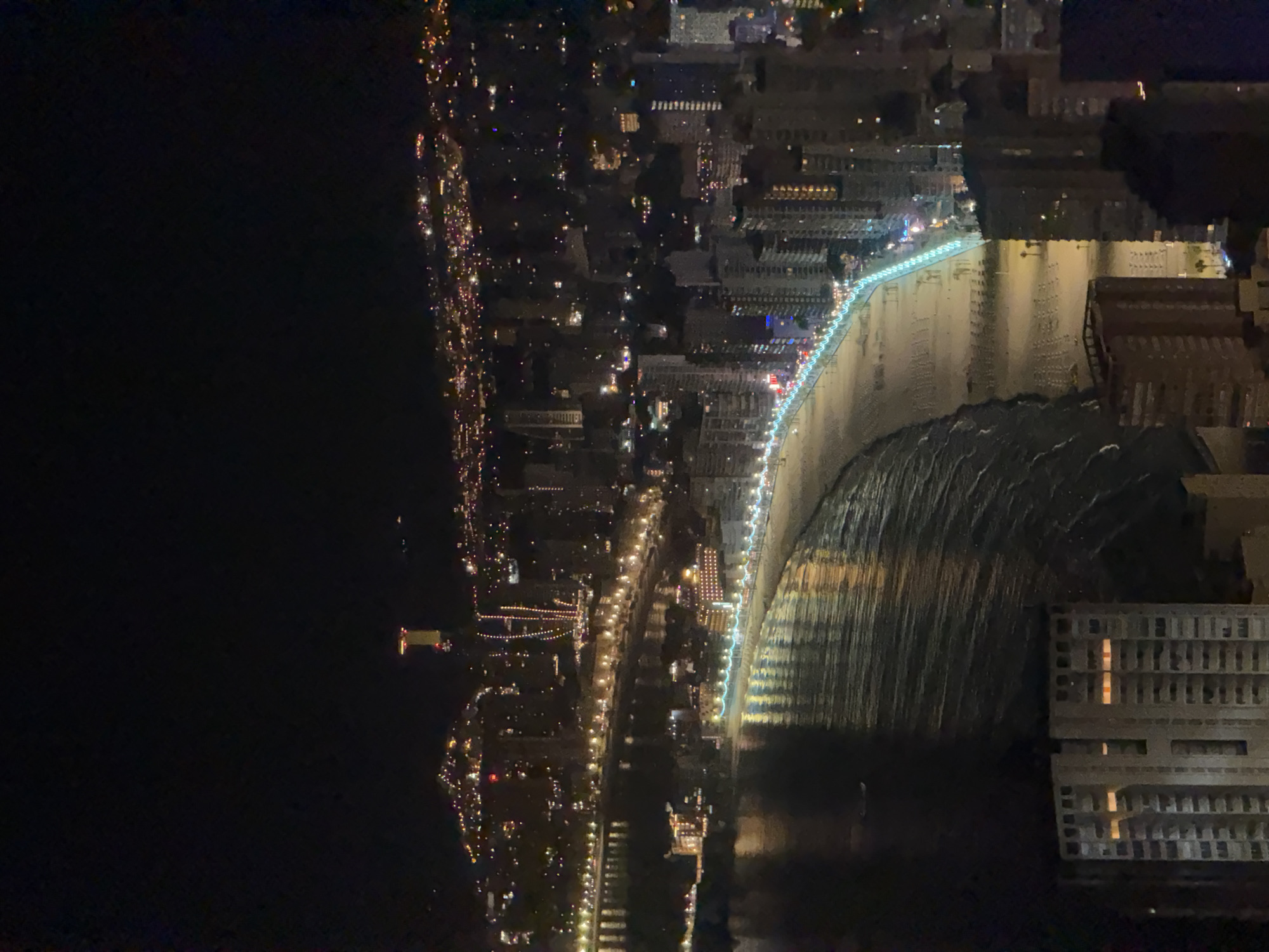 This is a nighttime aerial shot of the Benidorm coastline in Spain. The image showcases the vibrant city lights illuminating the beach and surrounding buildings. The focal point of the image is the crescent-shaped beach, which is lined with lights, creating a glowing path along the coastline. Numerous high-rise buildings are visible, with lights indicating occupied apartments or businesses. In the distance, the city extends up a hillside, dotted with more lights. The sea is dark, reflecting the city lights, and gentle waves are visible. A few boats are also present in the water. Some fireworks are visible in the upper portion of the image, suggesting a celebratory event.