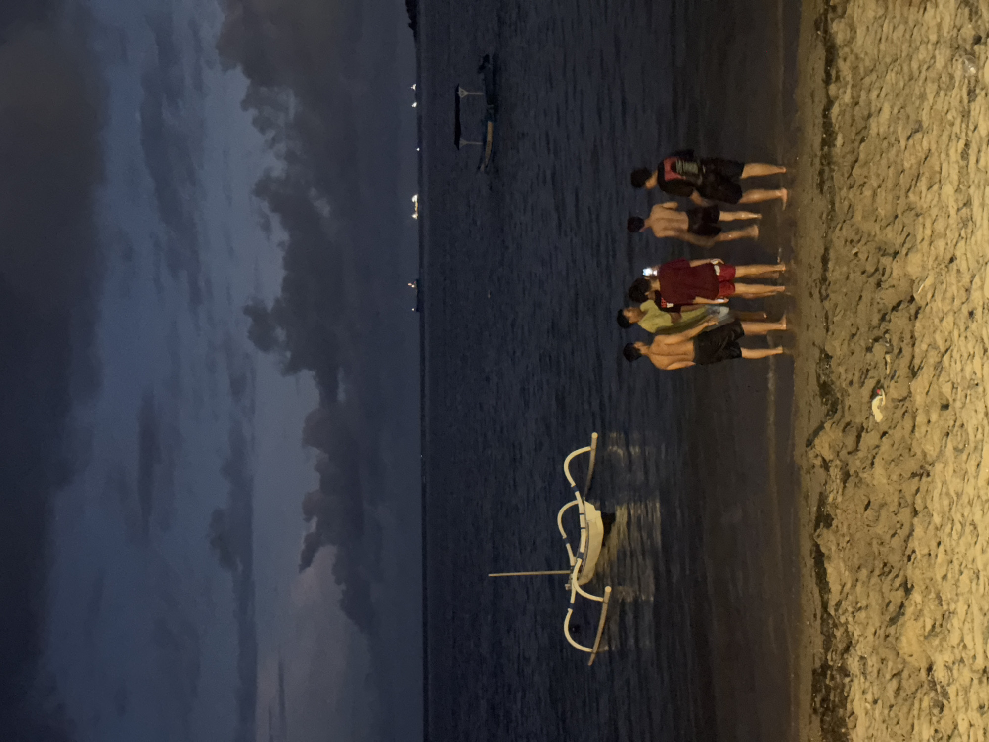 At dusk, a group of five males stand on a sandy beach at the water's edge. One holds a cell phone. The sea is calm and reflects the overcast sky. In the distance are lights that suggest a city. A boat is in the water. The scene is in Denpasar, Indonesia.