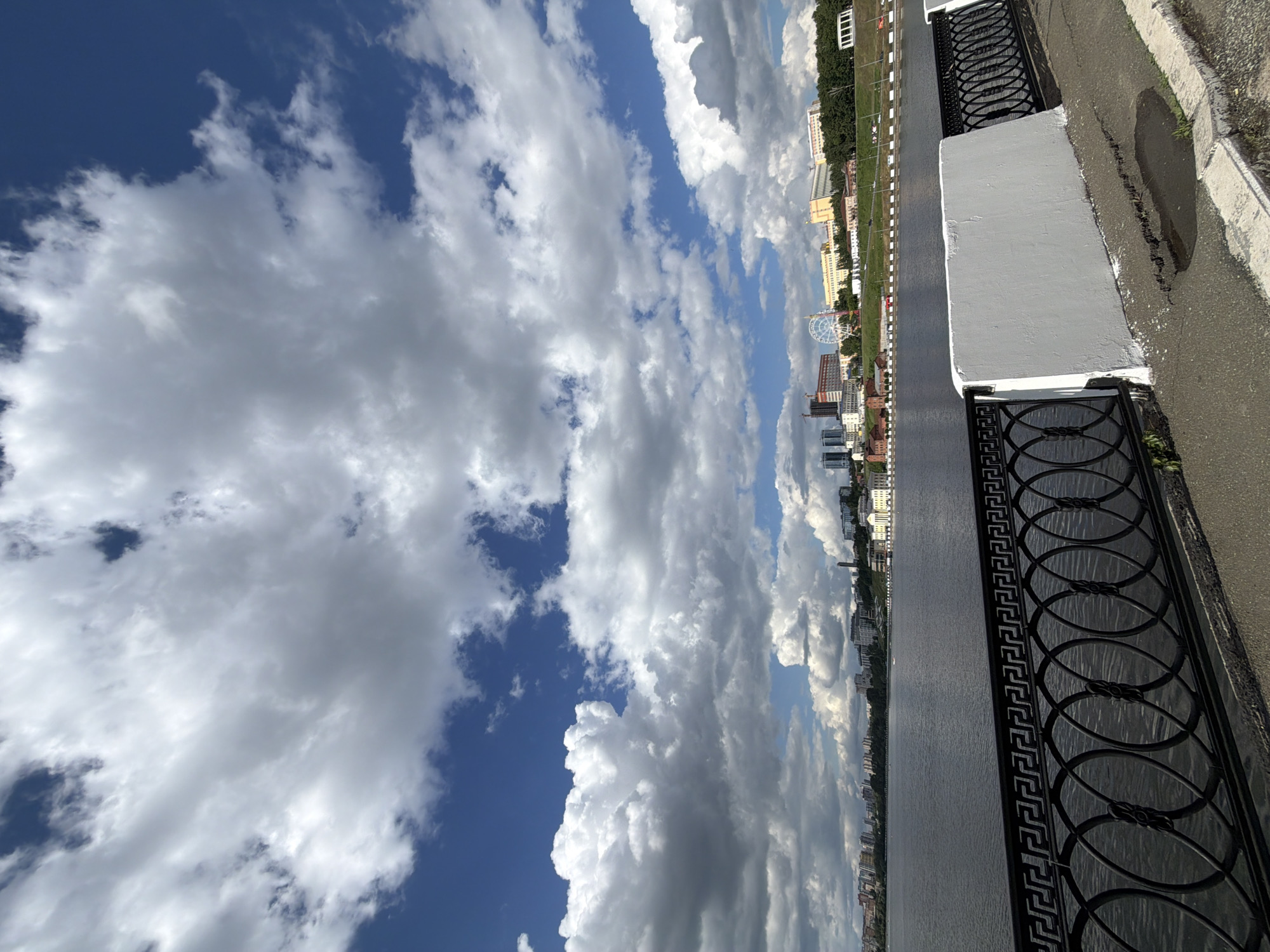 The image depicts a cityscape under a cloudy sky. The vantage point is from an elevated position, possibly a bridge or high bank, looking over water towards buildings and infrastructure. A black decorative metal railing runs along the bottom right edge of the image, bordering a light-colored concrete structure. Beyond that, a paved surface with some visible imperfections or puddles is present. On the other side of the water, various buildings of different heights and architectural styles are visible, including high-rise apartments and commercial structures. A ferris wheel can be seen in the distance. The sky is filled with dense, white clouds interspersed with patches of blue. The city is Izhevsk, Russia.