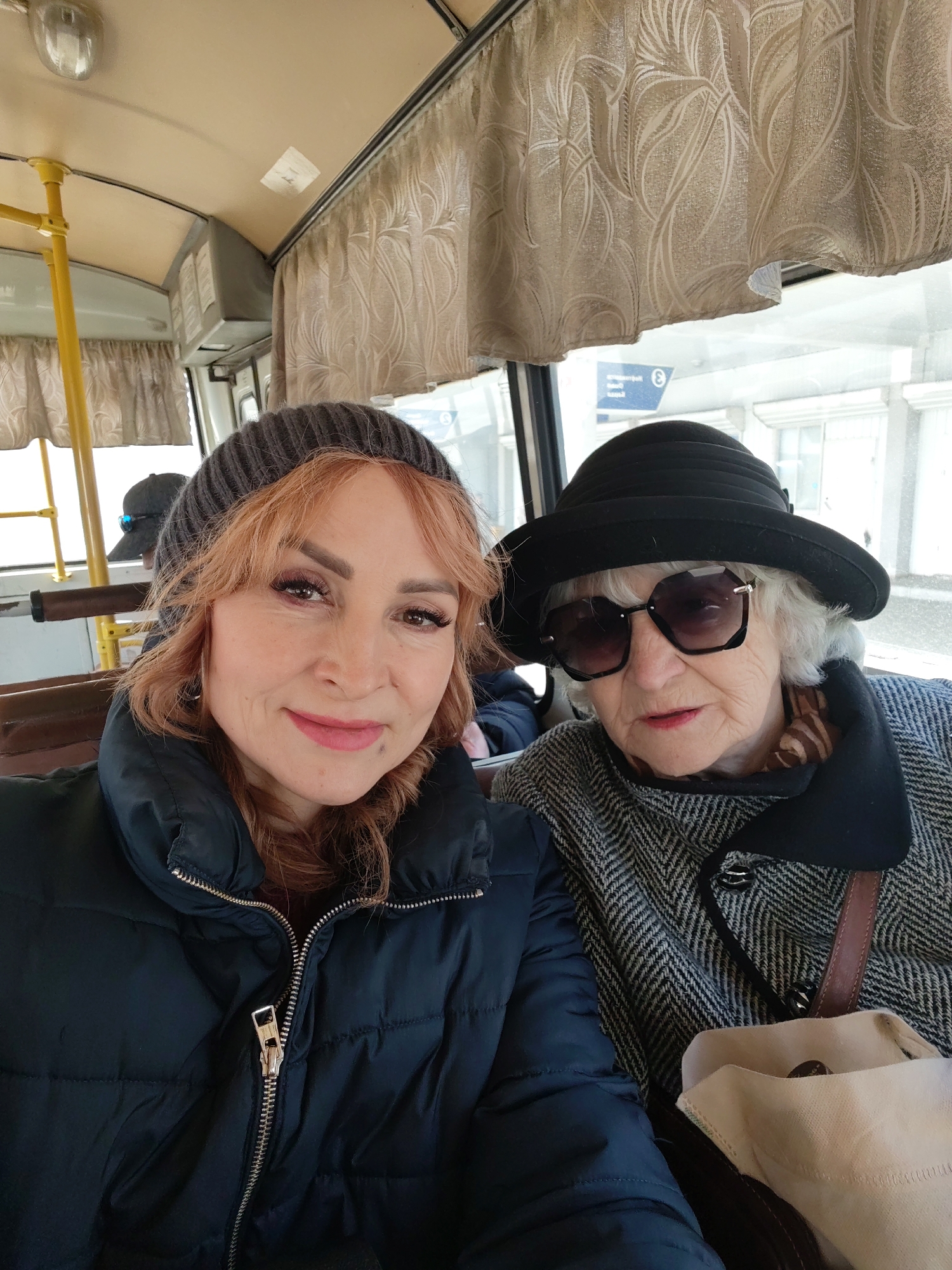 Two women are pictured inside a public transport vehicle, likely a bus or trolleybus, in Chaikovsky, Russia. The foreground features a younger woman with reddish-brown hair, wearing a grey knit beanie and a dark blue puffer jacket with a prominent silver zipper. She smiles gently towards the camera.
To her right, an older woman with white hair is visible, donning a black cloche hat and large, dark hexagonal sunglasses. She wears a black and white herringbone coat with a patterned scarf visible around her neck. Her expression is partially obscured by the sunglasses, but her mouth is slightly open. Next to her is a light-colored bag. Faintly visible in the background, further inside the vehicle, is another person wearing a dark baseball cap.
The interior of the vehicle suggests an older model, featuring light beige walls and ceiling, yellow metal poles for support, and decorative cream-colored curtains with a leaf pattern over the windows. An overhead light fixture and some indistinct grey panels are also visible. Through the window, a grey, possibly overcast sky and parts of an urban environment with buildings can be seen, indicating daytime. The warm attire of both women suggests cool weather. A blue sign with what appears to be "E 3" or similar is faintly visible outside the window on the right side. The image captures a casual moment, likely a selfie, of the two women riding together.
