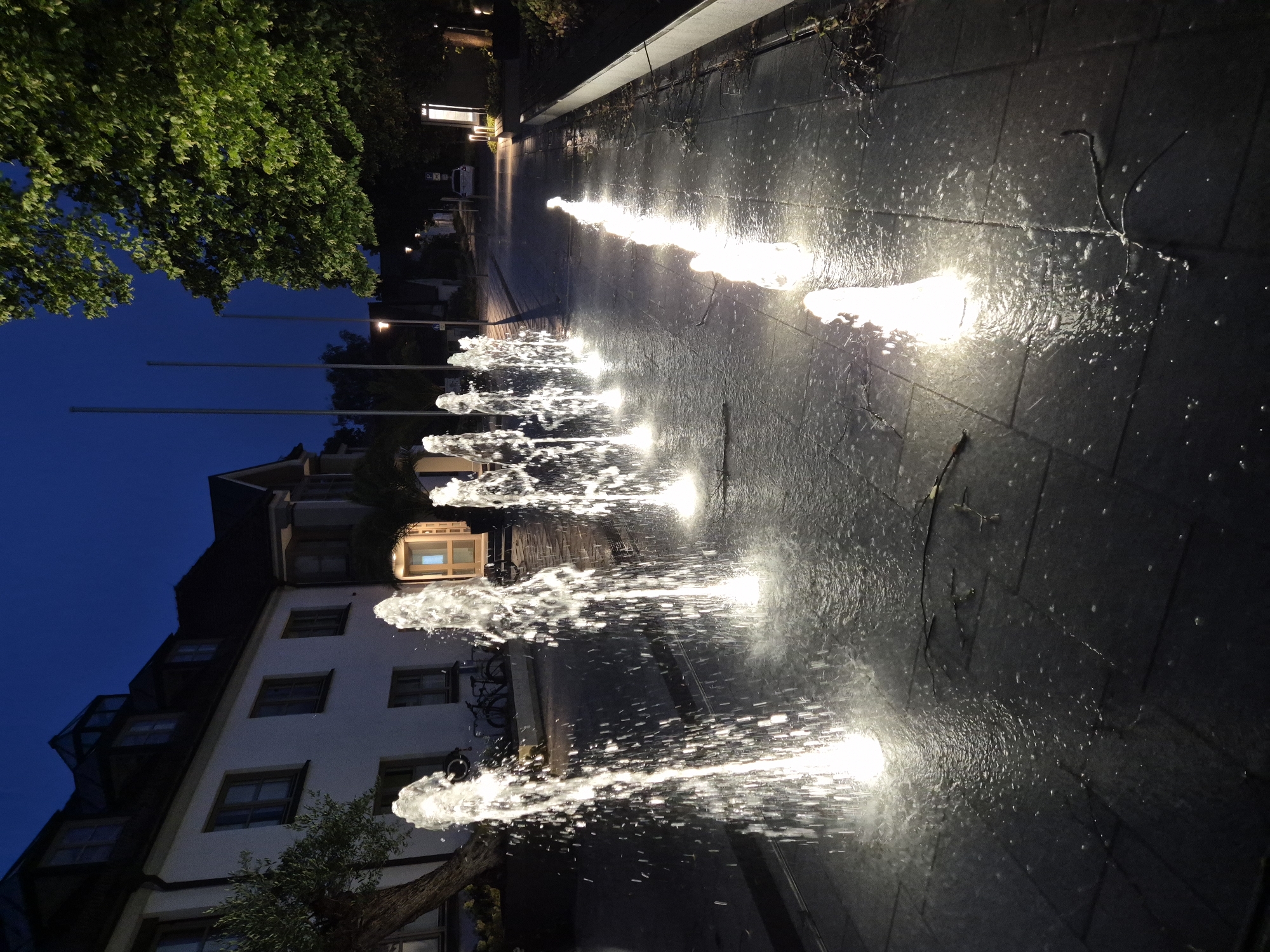 A row of illuminated water fountains are spraying water upwards from a paved surface at dusk. The fountains are arranged in two parallel lines on either side of a pathway that recedes into the distance. The water from the fountains is catching the light, creating a luminous effect. In the background, a building with multiple windows and a dark roof is visible on the left. On the right, a large tree with green leaves is illuminated by artificial light. A white car is parked further down the pathway. The overall scene suggests an outdoor public space, possibly in a town square or park, in Ergolding, Germany. The lighting indicates it is either early morning or late evening. There are no people visible in the image.