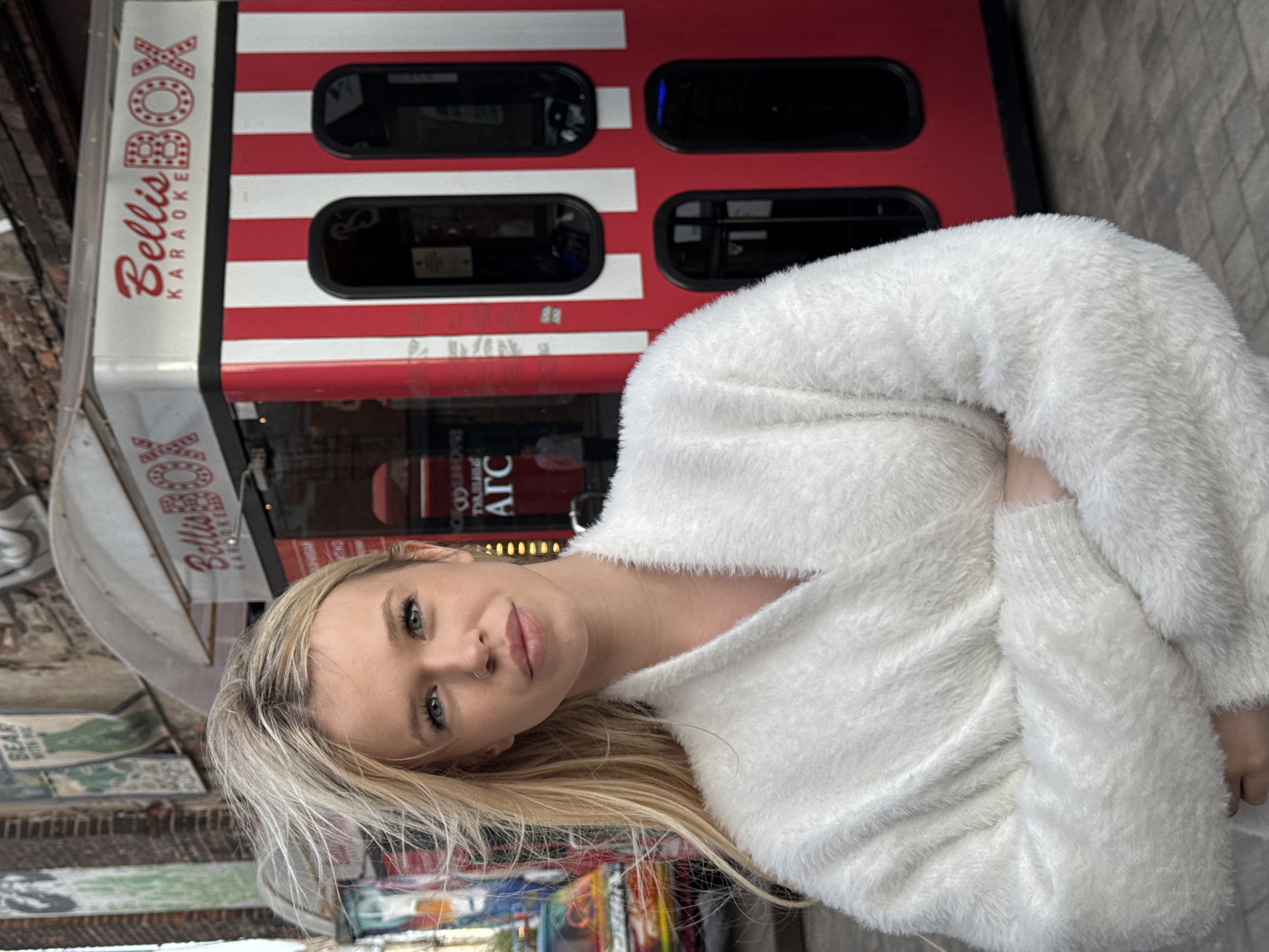 This media file depicts a young woman with blonde hair and light skin, wearing a fluffy white sweater. She has blue eyes and a nose ring. She is positioned in front of a red and white striped structure with "Bellis Karaoke" written on it in red script. The structure appears to be a booth or small venue. Several windows are visible on the structure, some with dark interiors. The setting seems to be outdoors, with a brick wall and other signs visible in the background, suggesting an urban environment. The lighting appears to be natural, implying daytime. The woman is looking towards the camera with a slight smile, conveying a relaxed and casual emotion. The overall impression is a candid shot of someone enjoying themselves in a public space. There is visible text on the structure stating "Bellis Karaoke" and "Bellis Box". There is also text on signs in the background, including "ATC". The location context provided is Saint Petersburg, Russia.