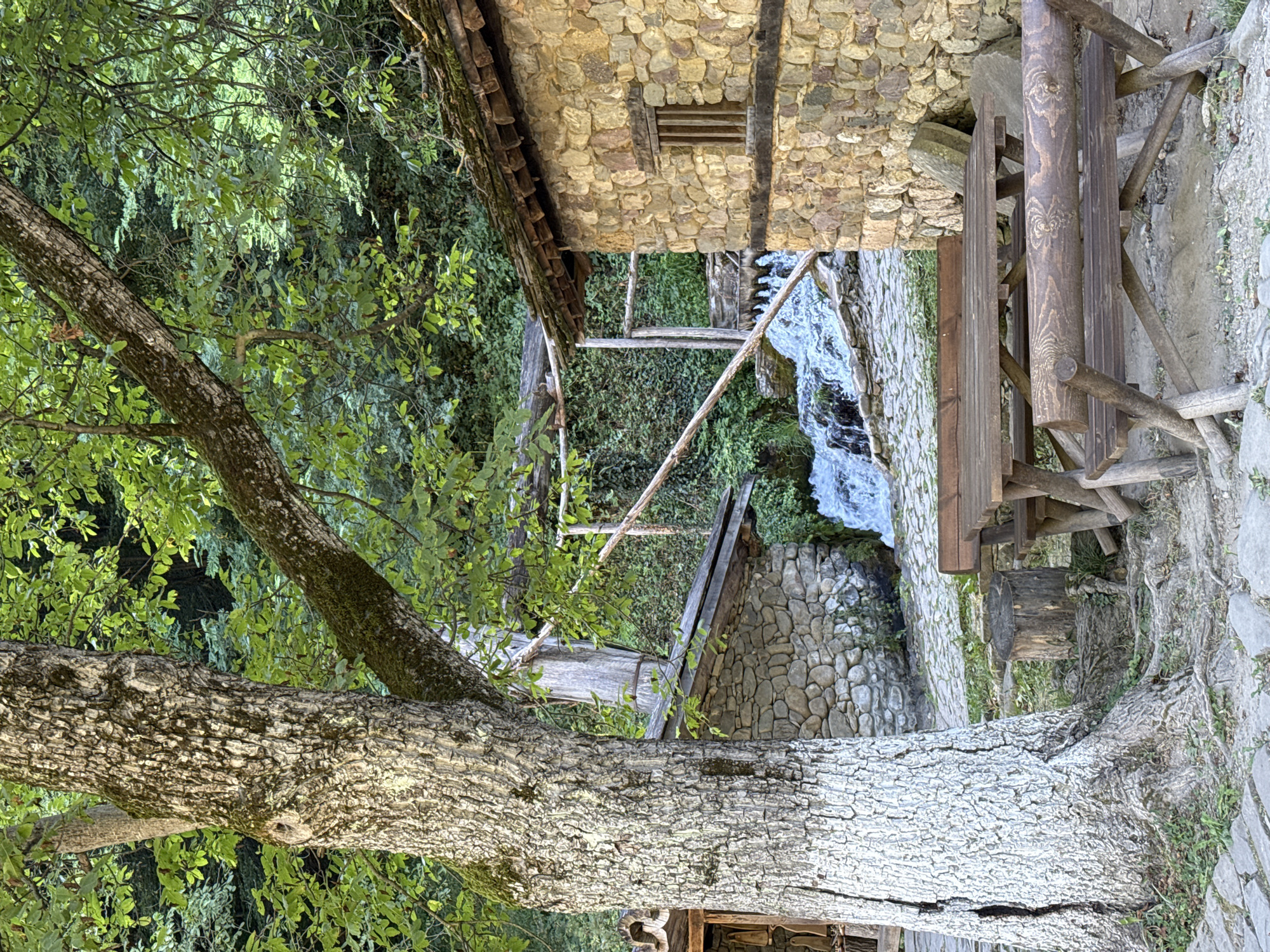 This is a photograph of a rustic outdoor seating area next to a small waterfall and stone buildings. In the foreground, a large tree with white-painted lower trunk stands on the left. A wooden picnic table with benches is positioned in front of it. The ground is paved with cobblestones and scattered with dirt and roots.
Behind the picnic table, a waterfall cascades down rocks. To the right of the waterfall, a stone building with a wooden-shingled roof and a small, barred window is visible. To the left of the waterfall, another stone building with a wooden roof is partially obscured by the large tree. Wooden structures and railings are also present around the waterfall and buildings. The background is filled with lush green foliage and trees, suggesting a natural, wooded environment.
This scene is located at the "House of Crafts" (Kъща на занаятите) in the village of Bozhentsi, near Gabrovo, Bulgaria. The area showcases traditional architecture and crafts. There are no people or animals visible in the image.