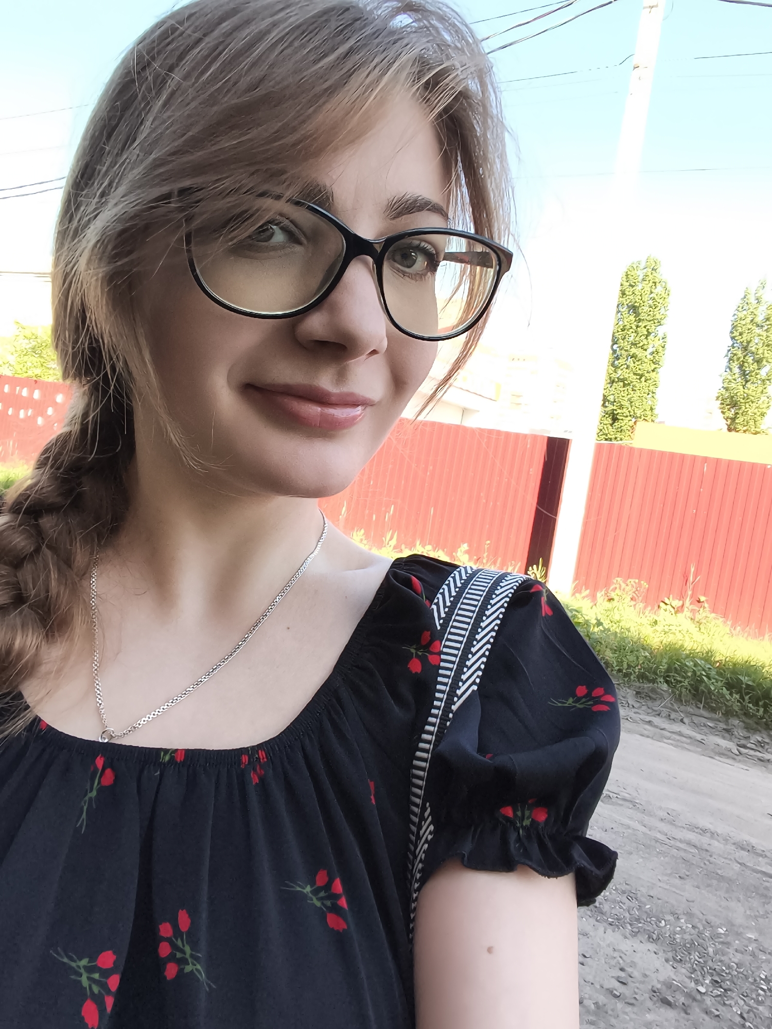 A young woman with light brown hair in a braid and dark-rimmed glasses takes a selfie outdoors. She is wearing a black dress with a floral pattern of red flowers and green leaves, and a silver necklace. The background shows a red fence, green foliage, and a dirt road. The sky is blue.