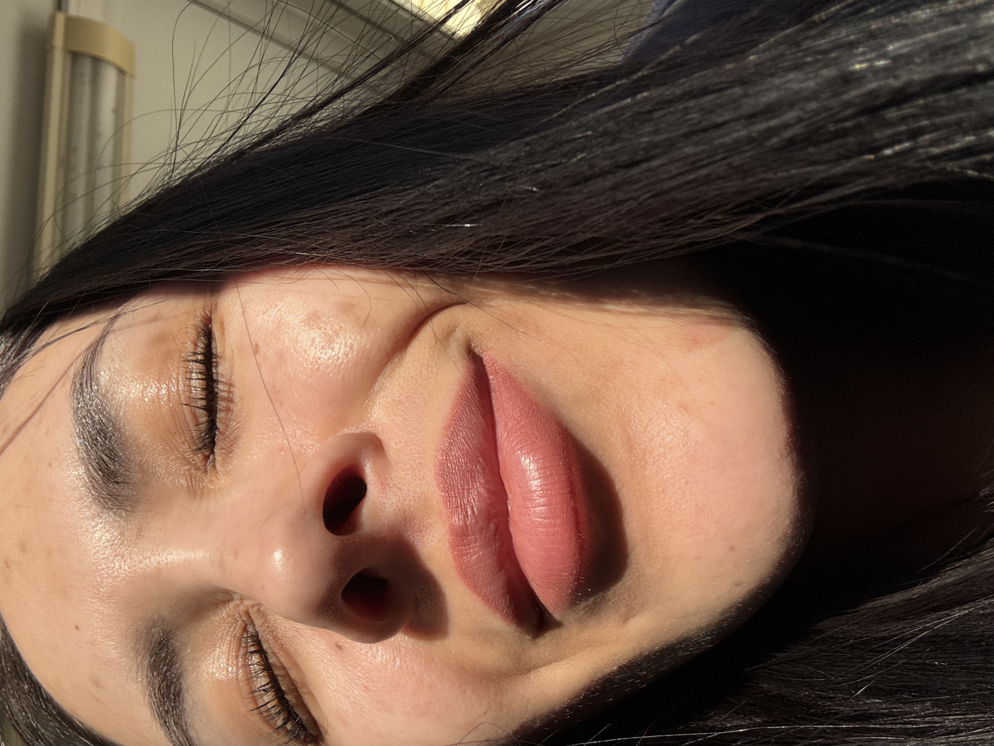 A close-up, indoor photograph, taken at an oblique angle, features a woman with closed eyes. Her face is predominantly bathed in sunlight, creating strong contrasts of light and shadow. She has dark hair, full lips with a rose-nude lipstick or gloss, and well-defined eyebrows. The background is indistinct, but hints of a window or light fixture are visible in the upper left corner.
The location in the city of Molodezhnyi, Russia, is not discernible from the image.