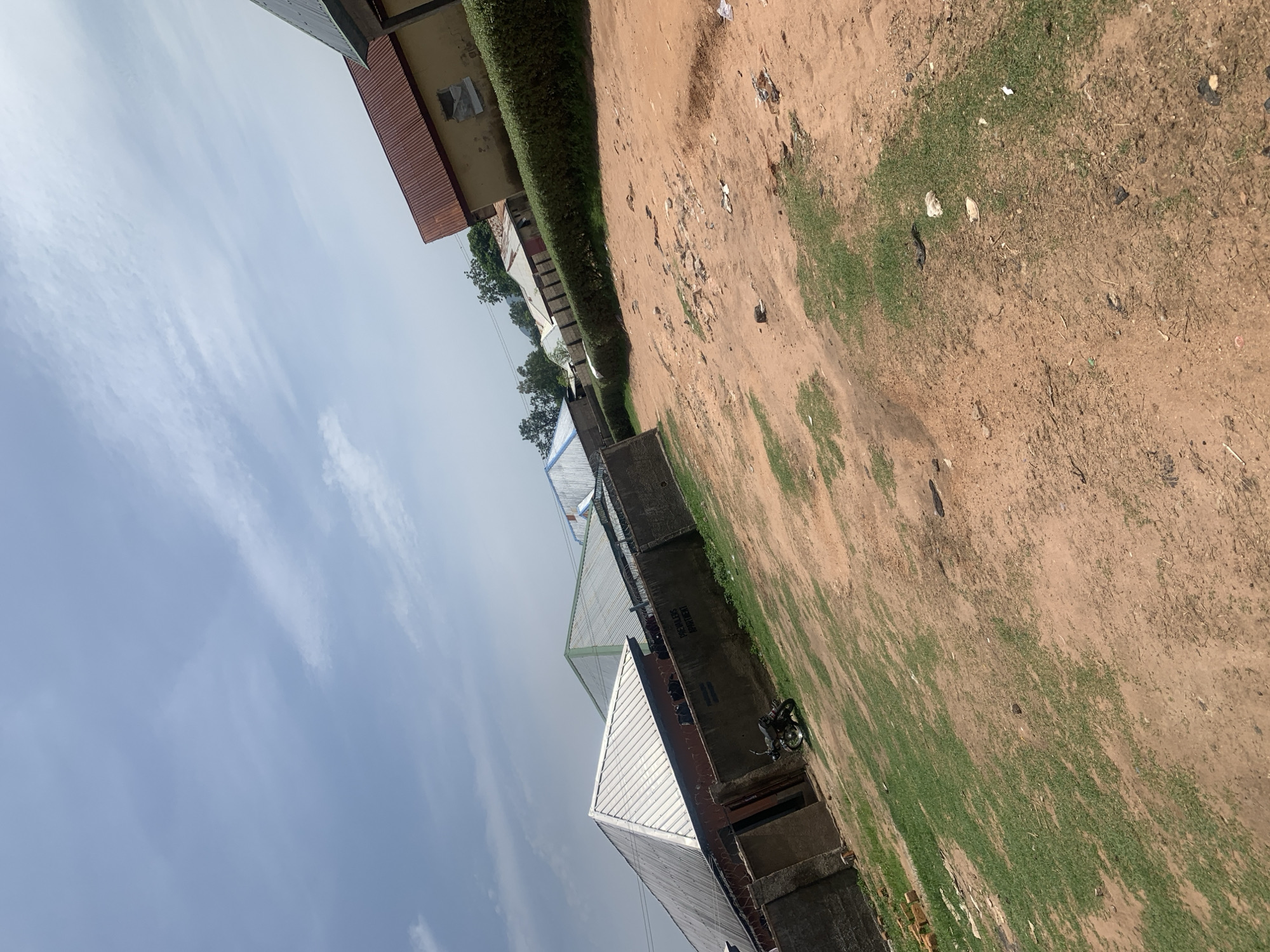 The image depicts a street scene in Jauro Yinu, Nigeria. The foreground is a dirt path with patches of dry grass and scattered debris. Several residential buildings with corrugated metal roofs are visible in the middle ground. One building on the left has a silver, pitched roof and a stone wall in front of it. A motorcycle is parked near this building. Further to the right, another building has a red, pitched roof and a yellow facade. A neatly trimmed hedge runs along the front of this building and the one next to it. In the background, more buildings and trees are visible under a cloudy sky. The overall impression is of a modestly developed urban or semi-urban area.