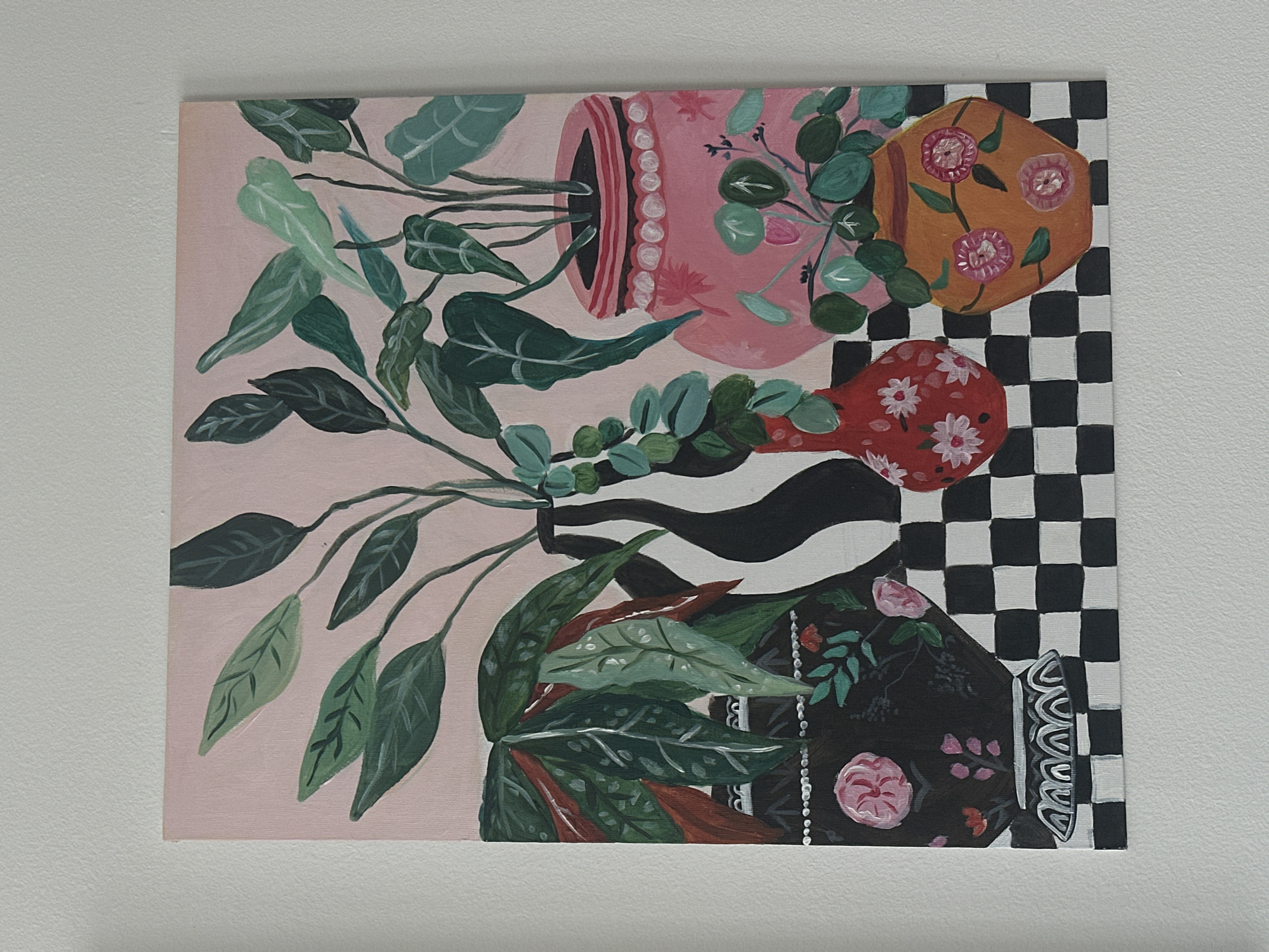 The image shows a painting of plants in pots. The painting depicts four pots of different colors and designs, arranged in a whimsical, slightly off-kilter manner. The pots are filled with various plants, characterized by broad, leafy foliage and delicate vines. The background consists of a combination of pink, black and white stripes, and a black and white checkerboard pattern. The painting is hanging on a plain, light-colored wall. The painting is not physically located in Novi Sad, Serbia; it is a depiction, a piece of art.