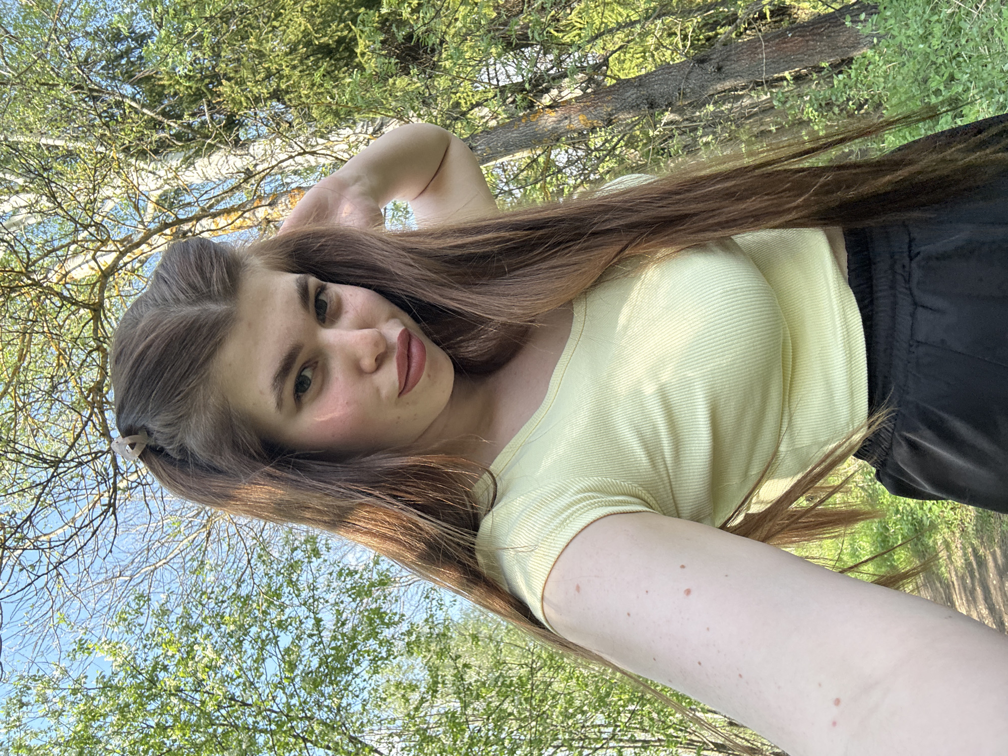 A young woman is posing for a selfie outdoors. She has long brown hair, is wearing a light yellow short-sleeved shirt, and black pants or shorts. She is standing or positioned with trees and foliage as the backdrop, with the sky visible through the branches. There is no indication that the image was taken in the city of Шевлягино, Russia.